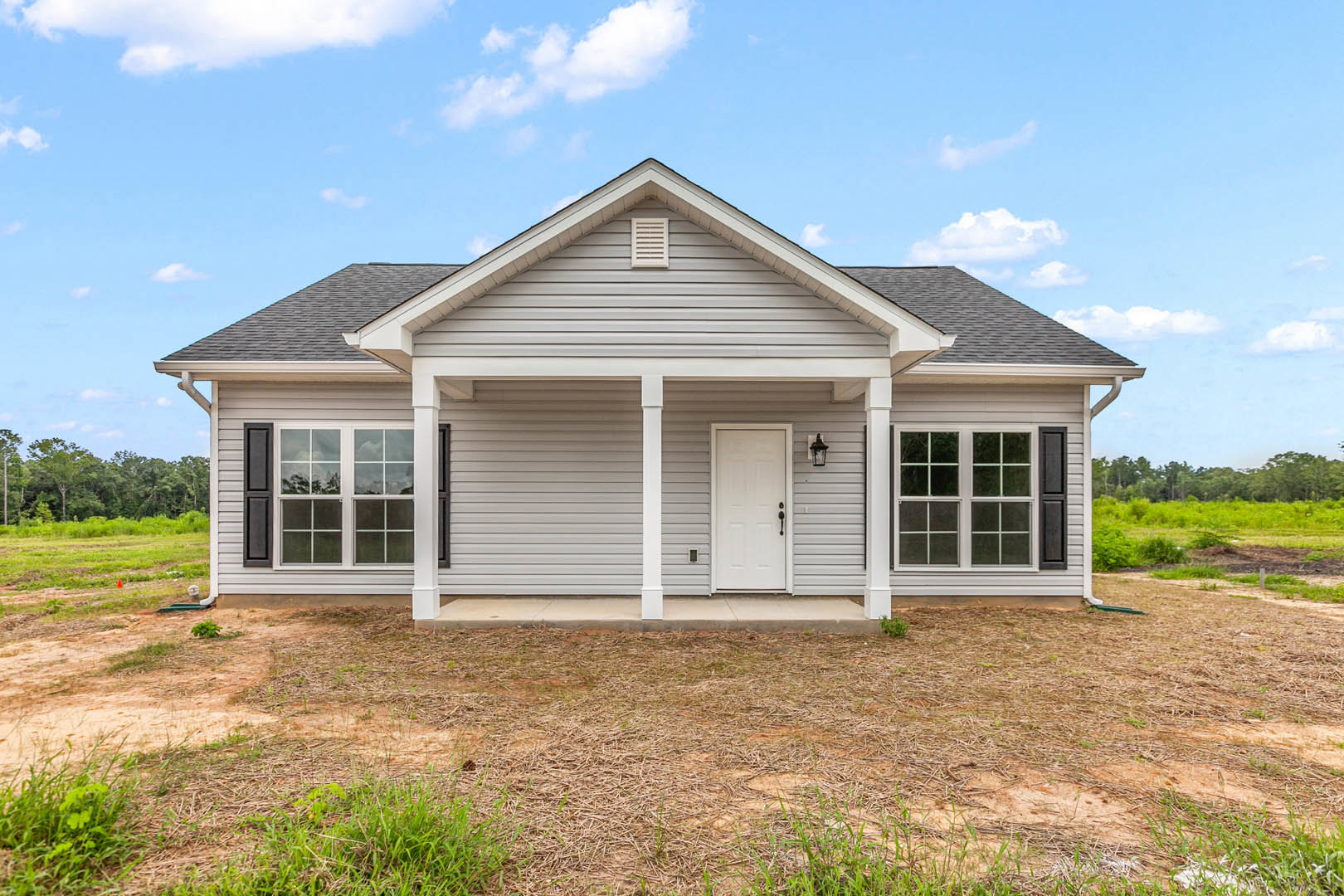 Grey siding house with white trim, black handle on white front door, white vent, windows, grassy yard, white support pole, shingled roof, cloudy sky in background