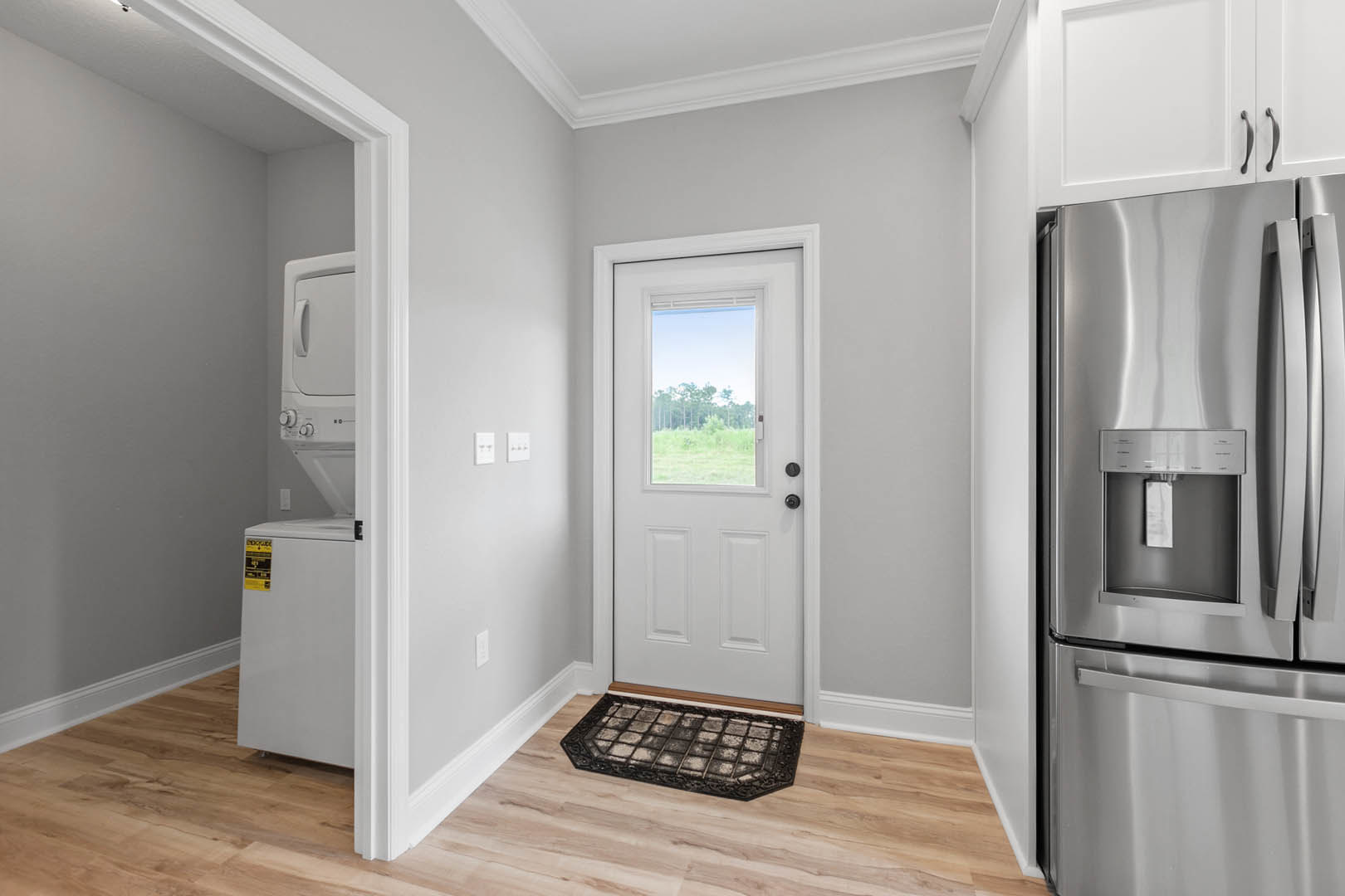 White paneled door with glass window, adjacent to white wall and yellow sign, refrigerator and cabinetry visible in kitchen, wood flooring with dirty metal grate, window