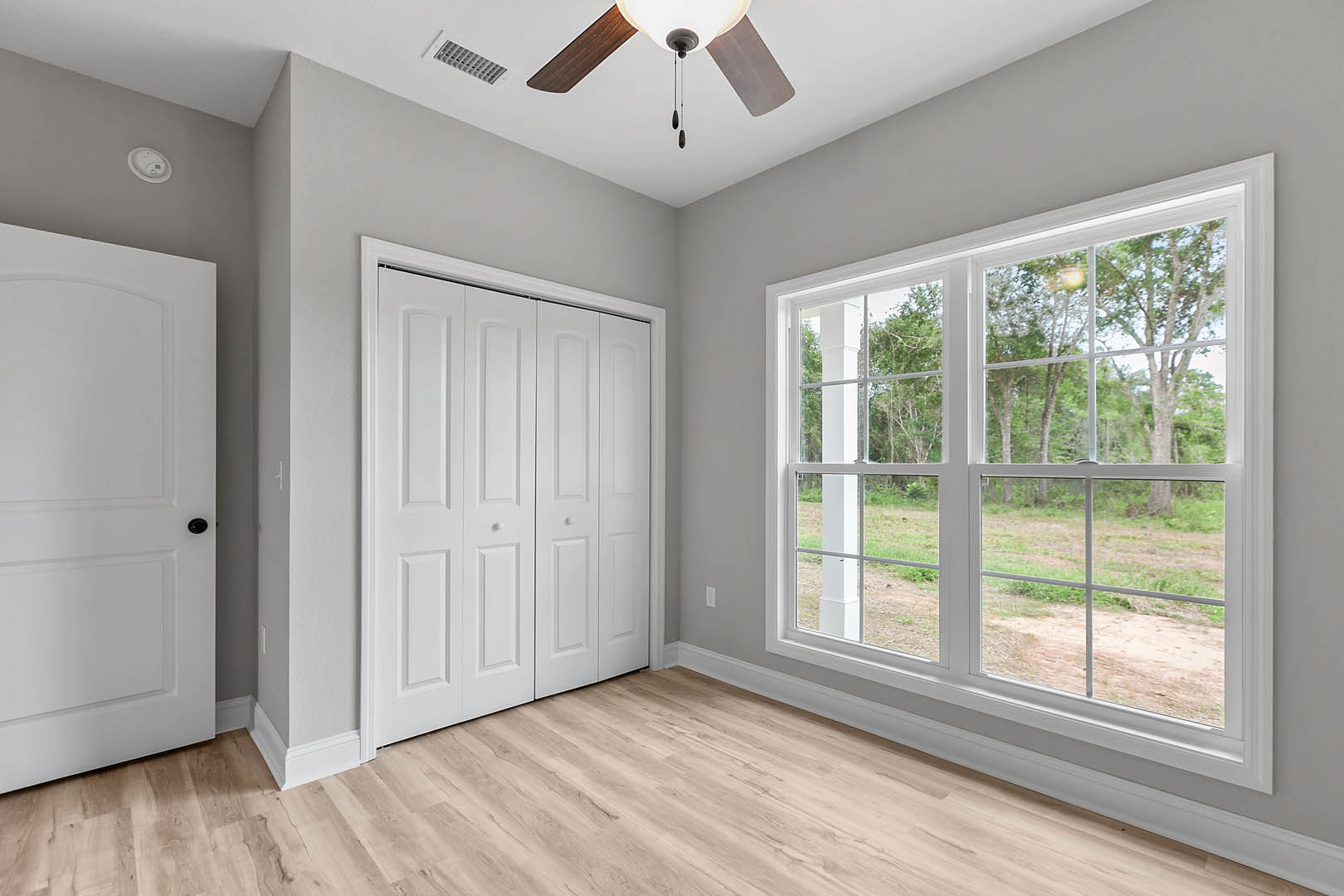 Bedroom with wood flooring, white baseboards, white closet doors, ceiling fan, and window overlooking trees