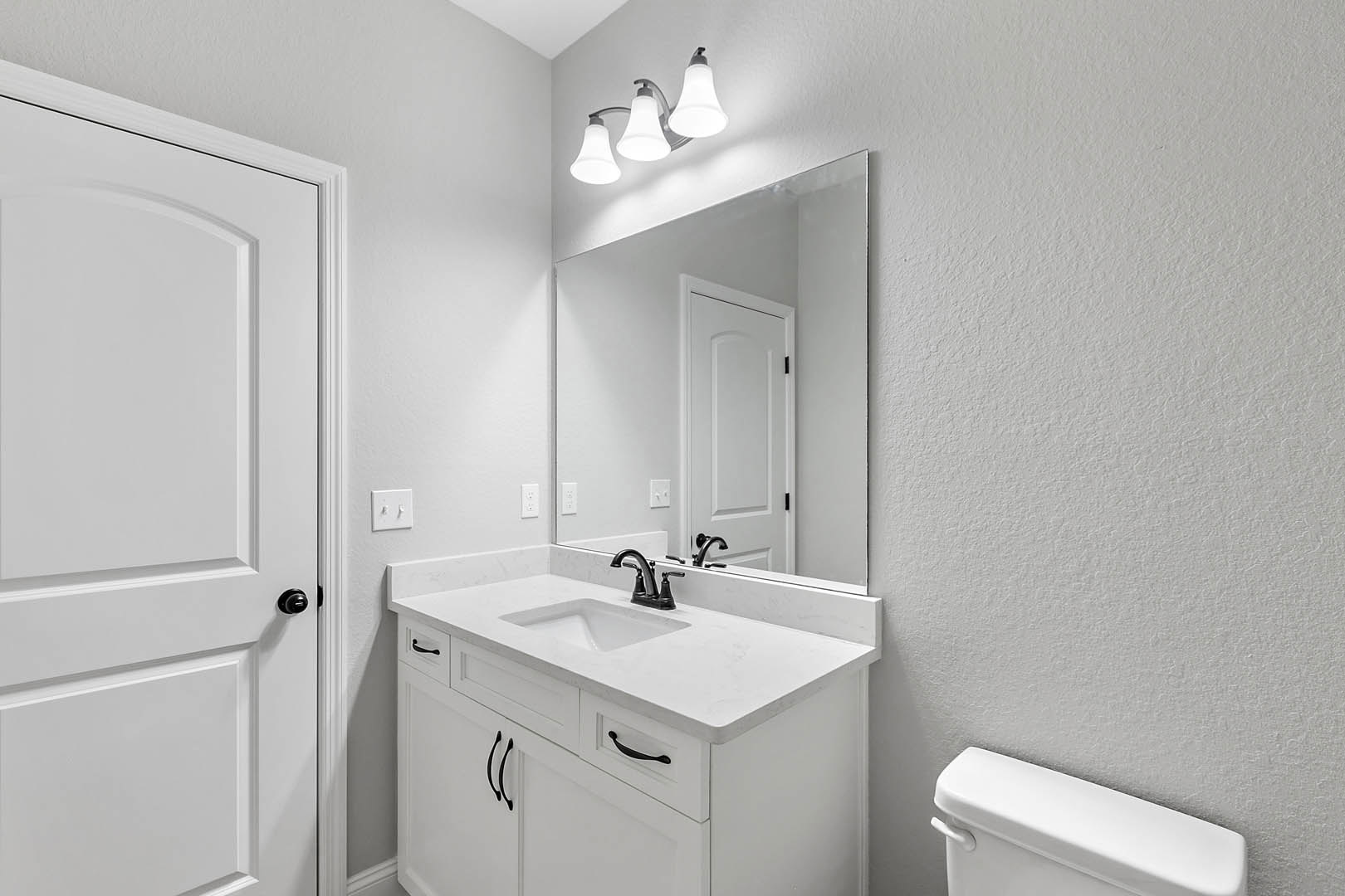 White bathroom with rectangular sink featuring black handles, adjacent toilet, white tile walls, three-bulb light fixture above mirror, white door, and dual light switch.