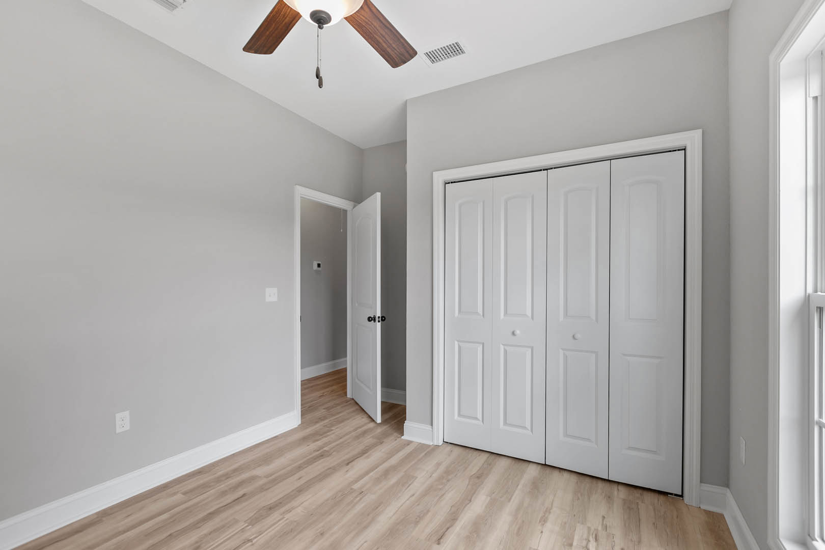 Wood floor with white baseboard trim, white paneled doors featuring black knobs, ceiling fan with light fixture, and air vent on smooth plaster wall