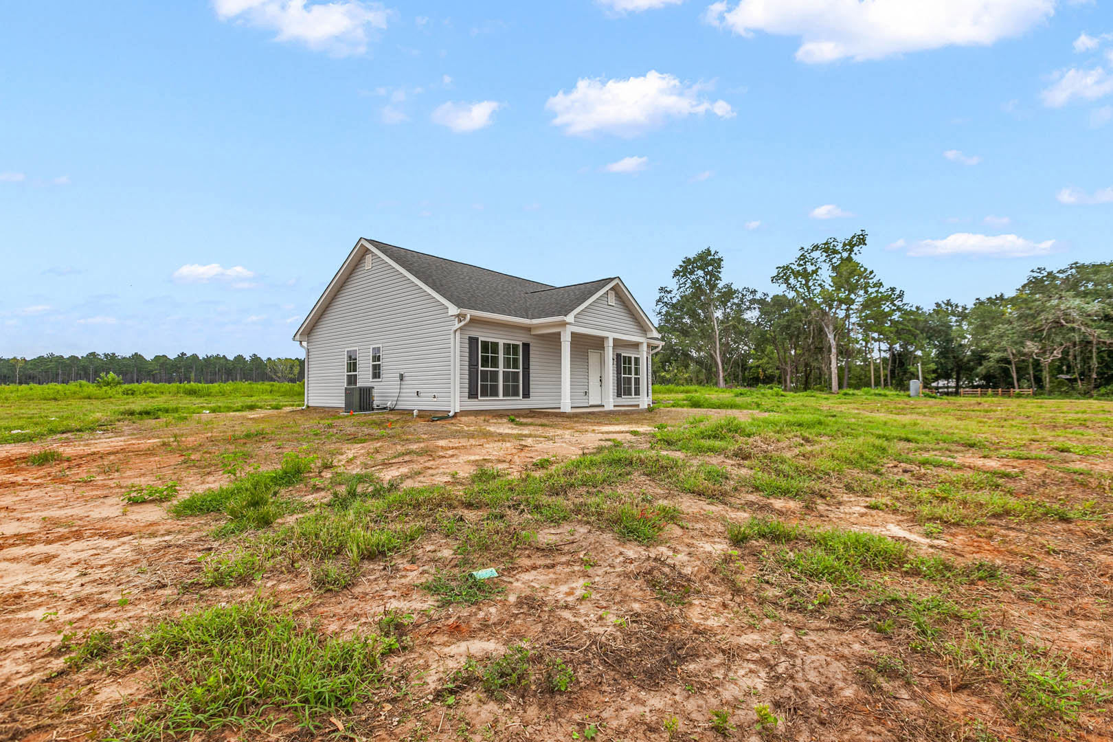 Modern farmhouse with white roof and white-trimmed windows, surrounded by grassy field, patch of dirt, and scattered trees under blue sky with white clouds
