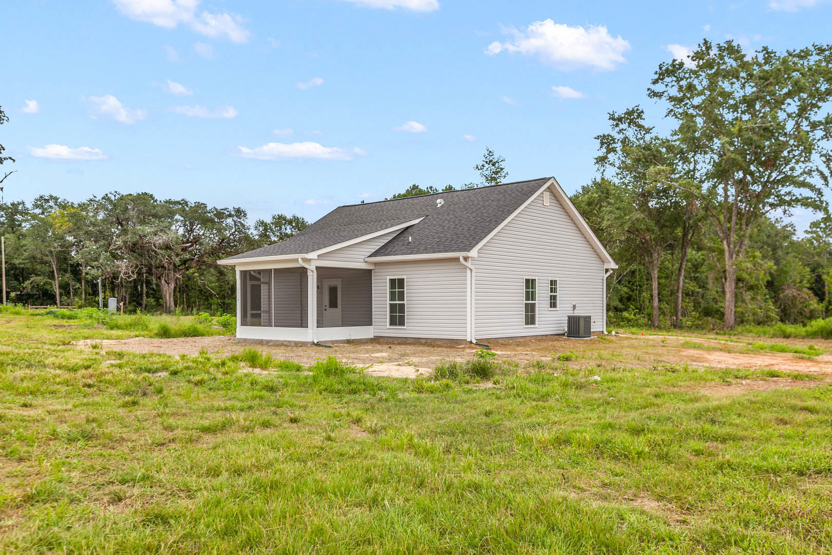 Two-story farmhouse with white siding, front porch, and side porch, surrounded by grass lawn and mature leafy trees under a partly cloudy sky