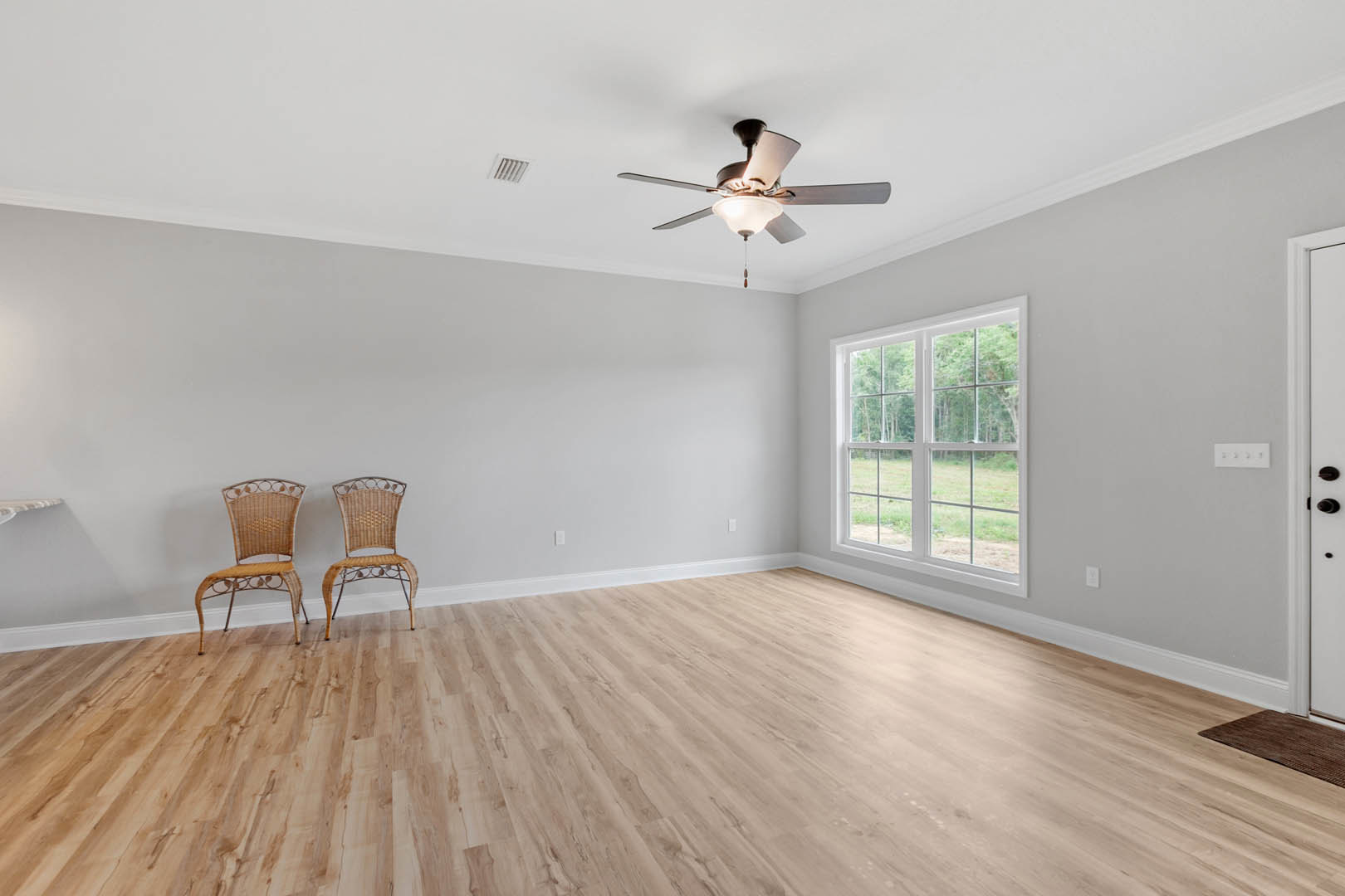 Ceiling fan with light fixture above two wicker chairs with metal frames, hardwood floor, large window overlooking trees, plaster walls