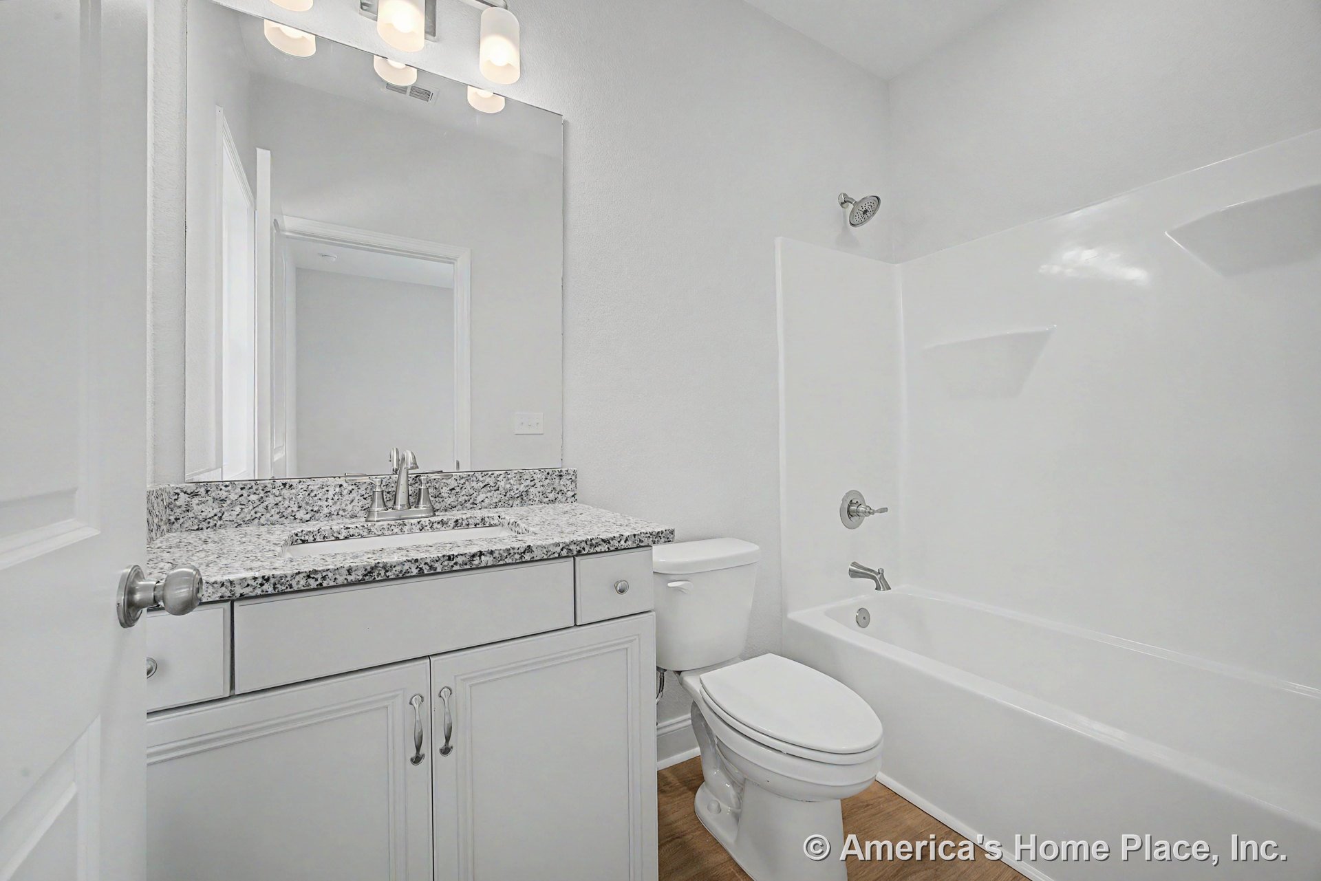 Granite countertop vanity with single chrome faucet, white shaker cabinets, large wall mirror beneath three-light fixture, tub-shower combination with integrated shelves, wood-look