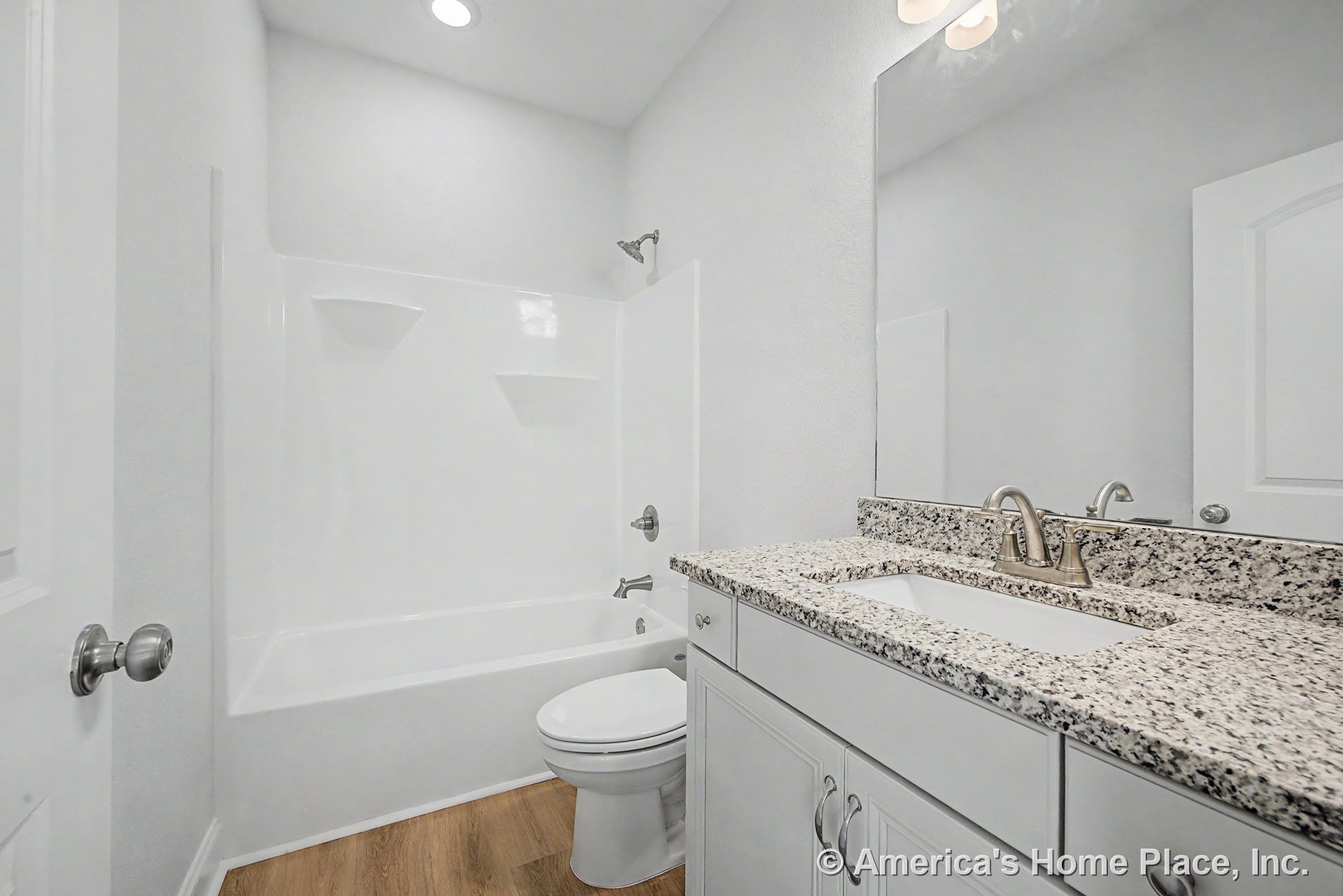 Granite vanity countertop with white shaker cabinets, built-in bathtub-shower combo, large wall mirror, recessed ceiling light, wood-look vinyl flooring, white trim, chrome faucet