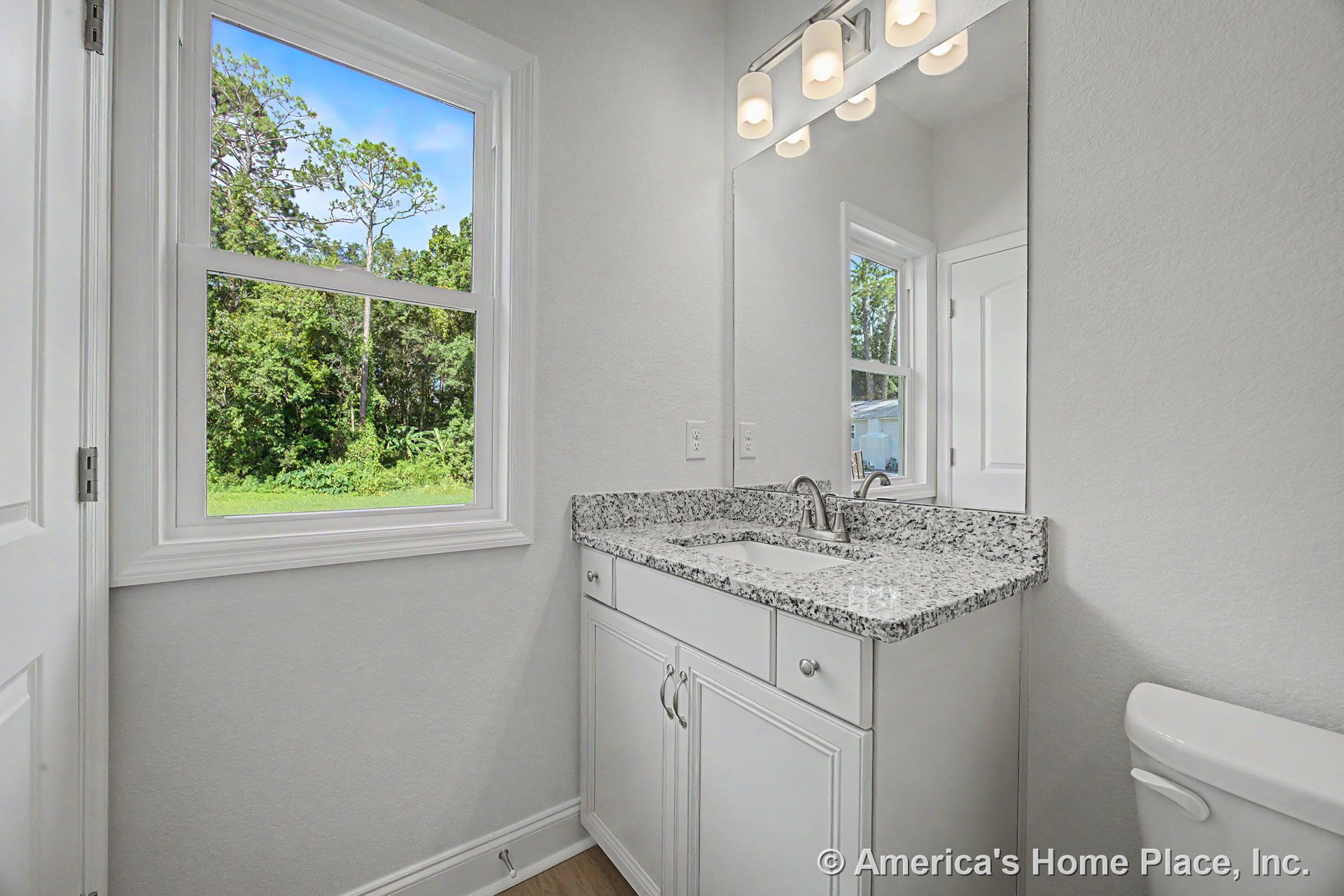 White shaker vanity with granite countertop, large wall mirror above, three-light fixture, double-hung window with trim, neutral wall paint, chrome faucet and undermount sink.