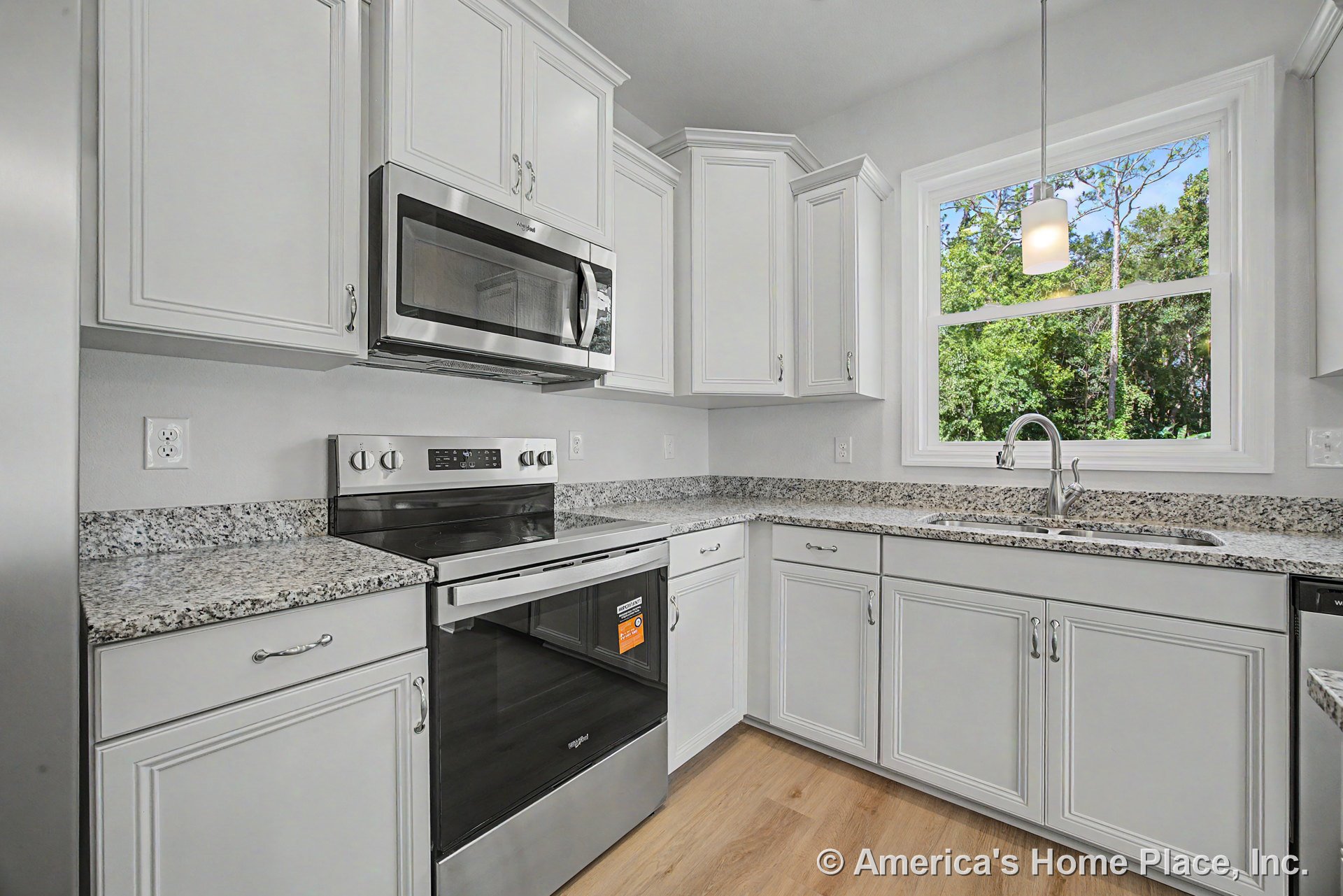 White upper and lower cabinets with granite countertops, stainless steel oven and microwave, large window above sink, pendant light fixture, wood-look flooring.