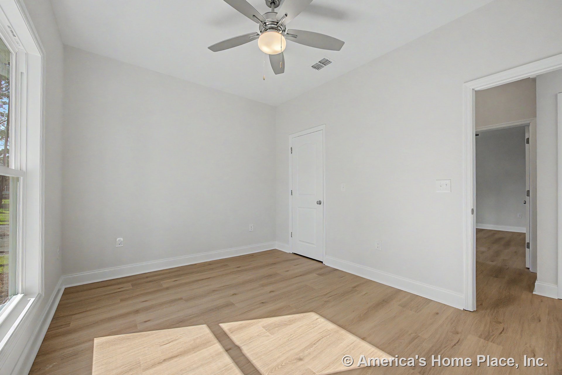 Bedroom with light wood plank flooring, white trim and baseboards, ceiling fan with integrated light, large window allowing natural light, white interior doors, and neutral wall