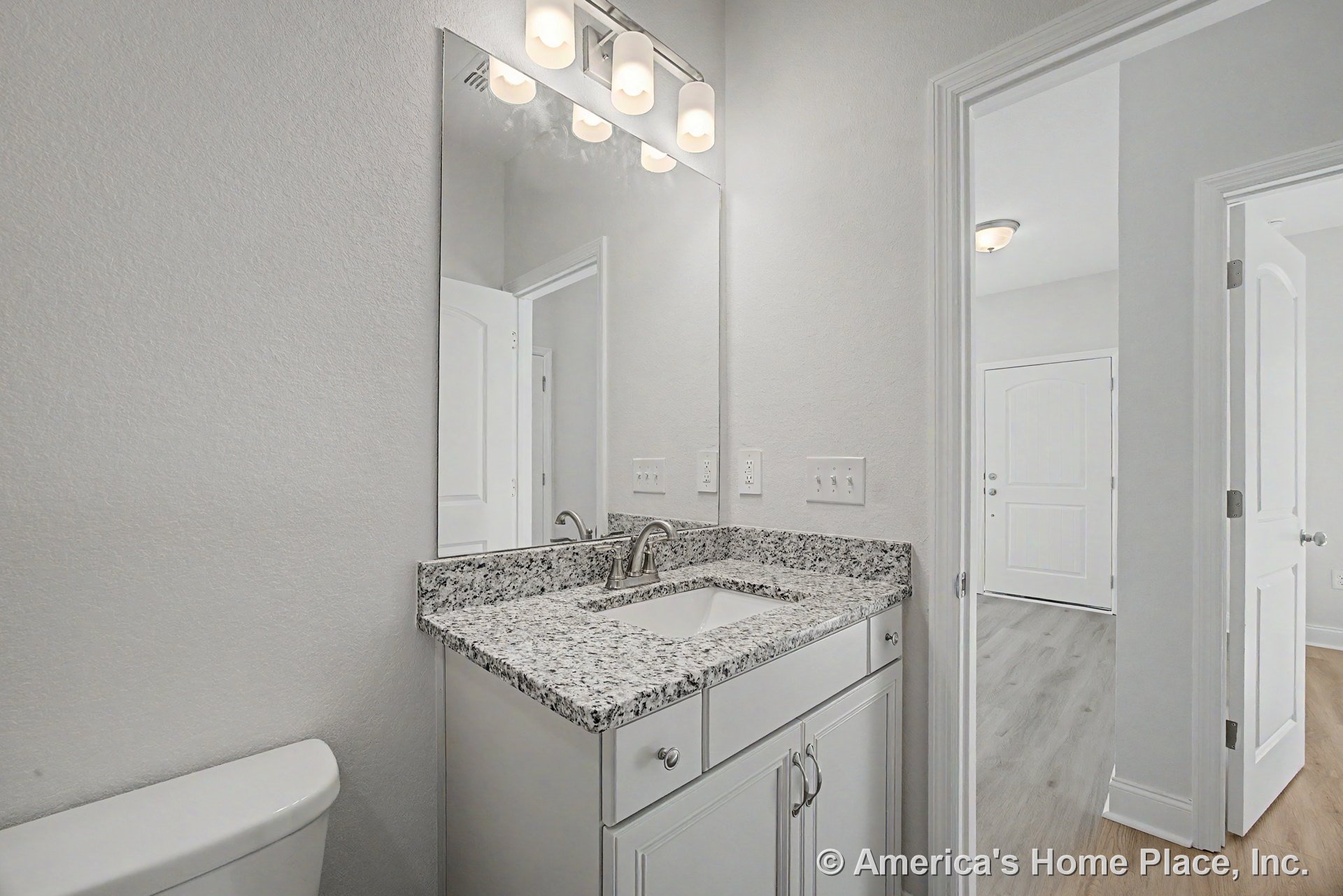 Granite countertop vanity with rectangular undermount sink, three-light fixture above expansive wall mirror, white cabinetry, neutral wall color, wood-look flooring visible in