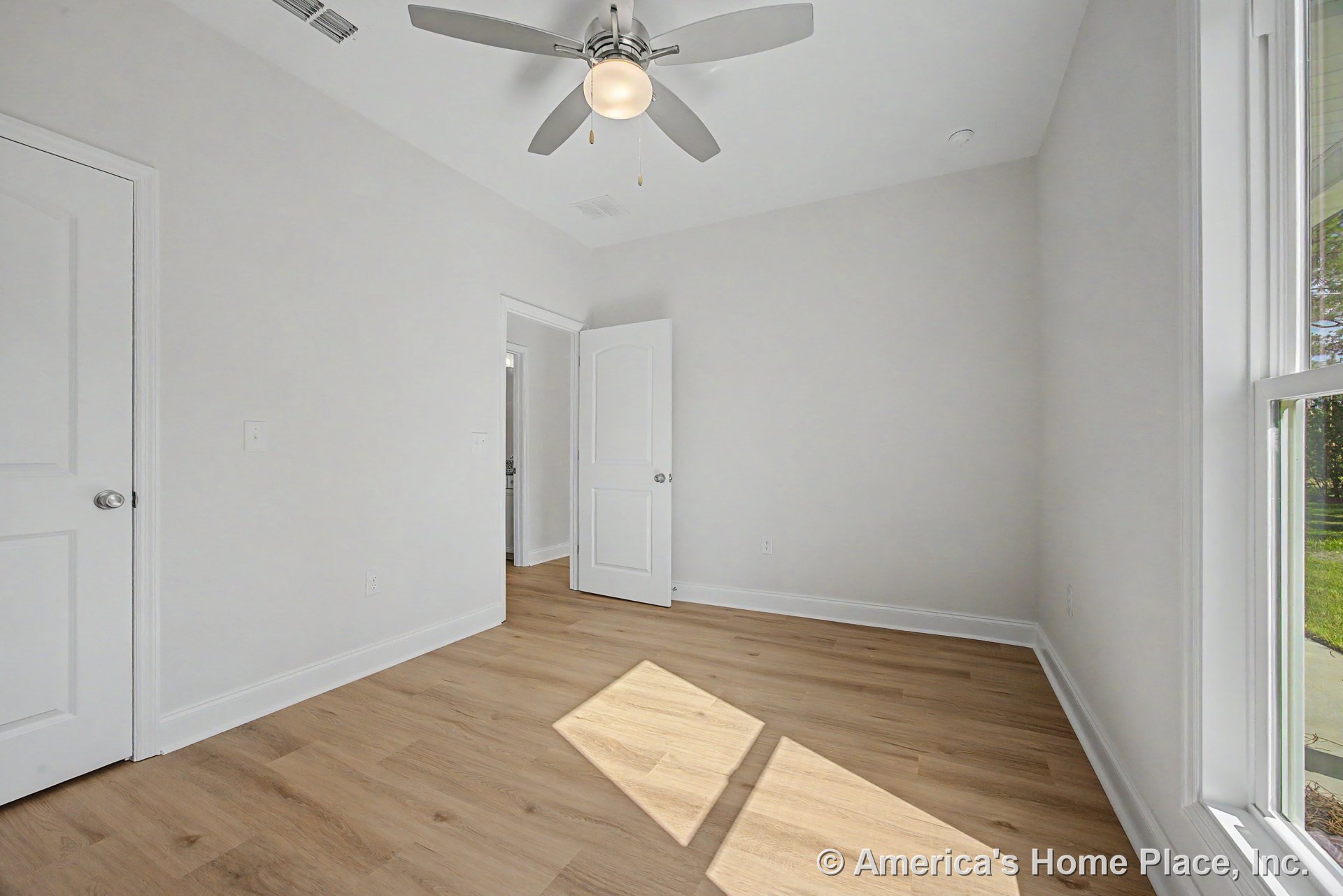 Light wood plank flooring, white baseboards and trim, large window with white frame, ceiling fan with integrated light fixture, white paneled doors, and neutral painted walls in a