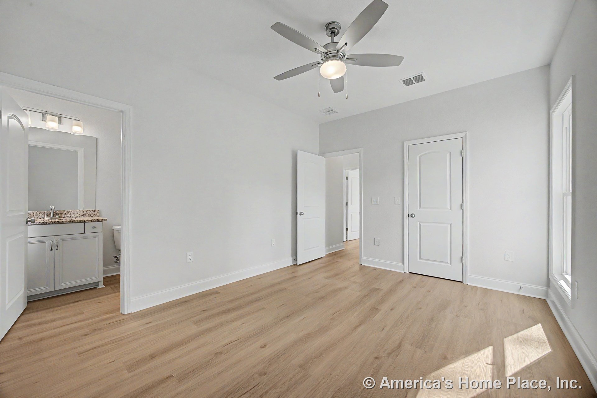 Bedroom with light wood plank flooring, white baseboards and trim, ceiling fan with integrated light fixture, double panel doors, large window with white casing, and bathroom
