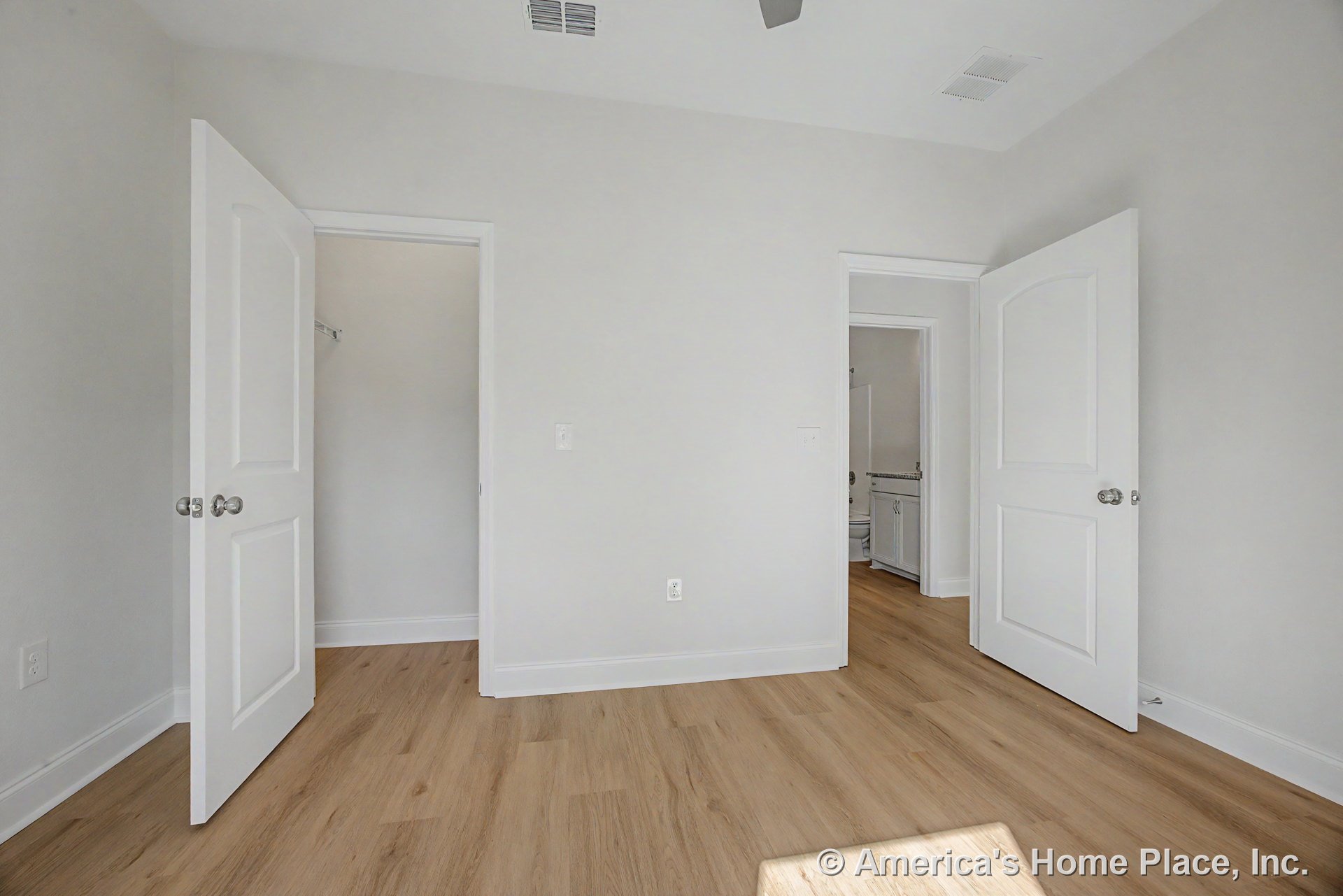 Bedroom with light wood plank flooring, white baseboards and trim, two white paneled doors providing access to closet and bathroom, neutral wall color, ceiling vent, and door