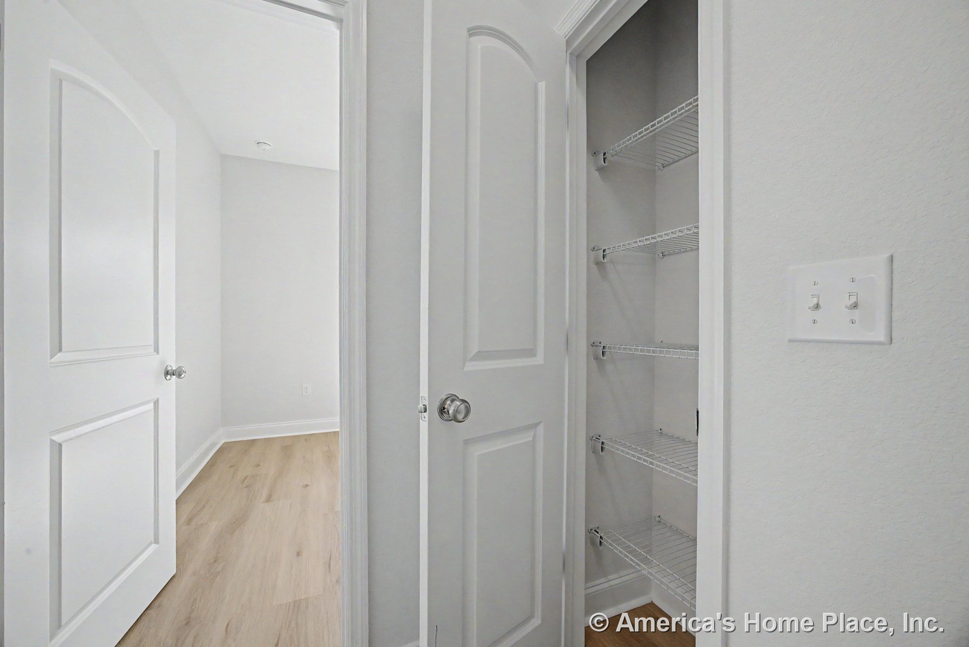 Hallway with white paneled closet doors, built-in wire shelving, light wood flooring, white baseboards and trim, double light switch, and neutral wall color.