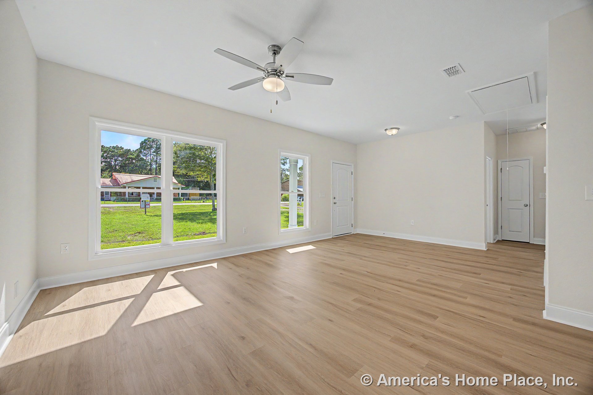 Spacious living room with large double windows, light wood plank flooring, white trim and baseboards, ceiling fan with built-in lighting, paneled entry door, and neutral wall