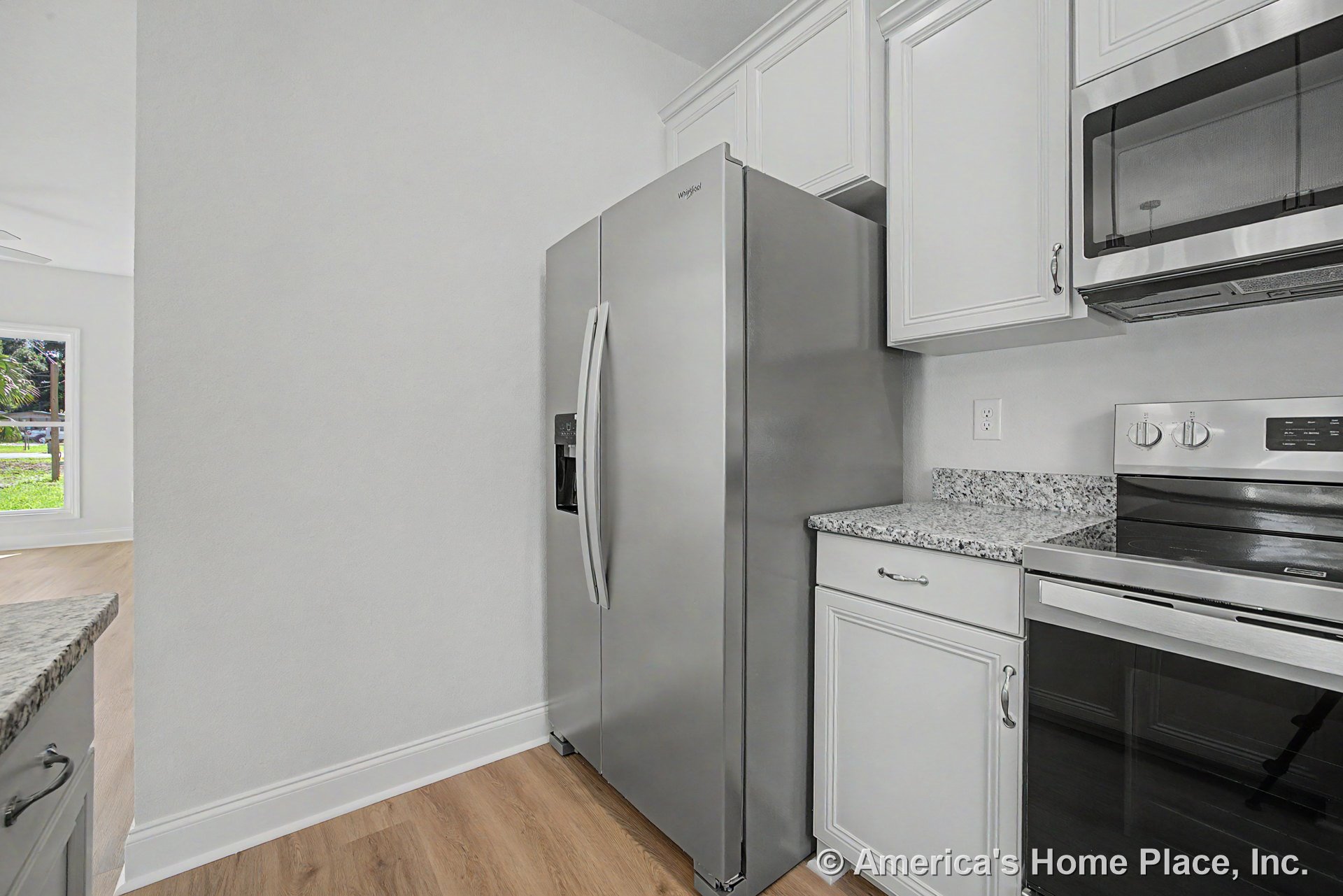 White upper and lower cabinets with granite countertops, stainless steel side-by-side refrigerator, oven, and microwave; light wood flooring and a window with decorative trim