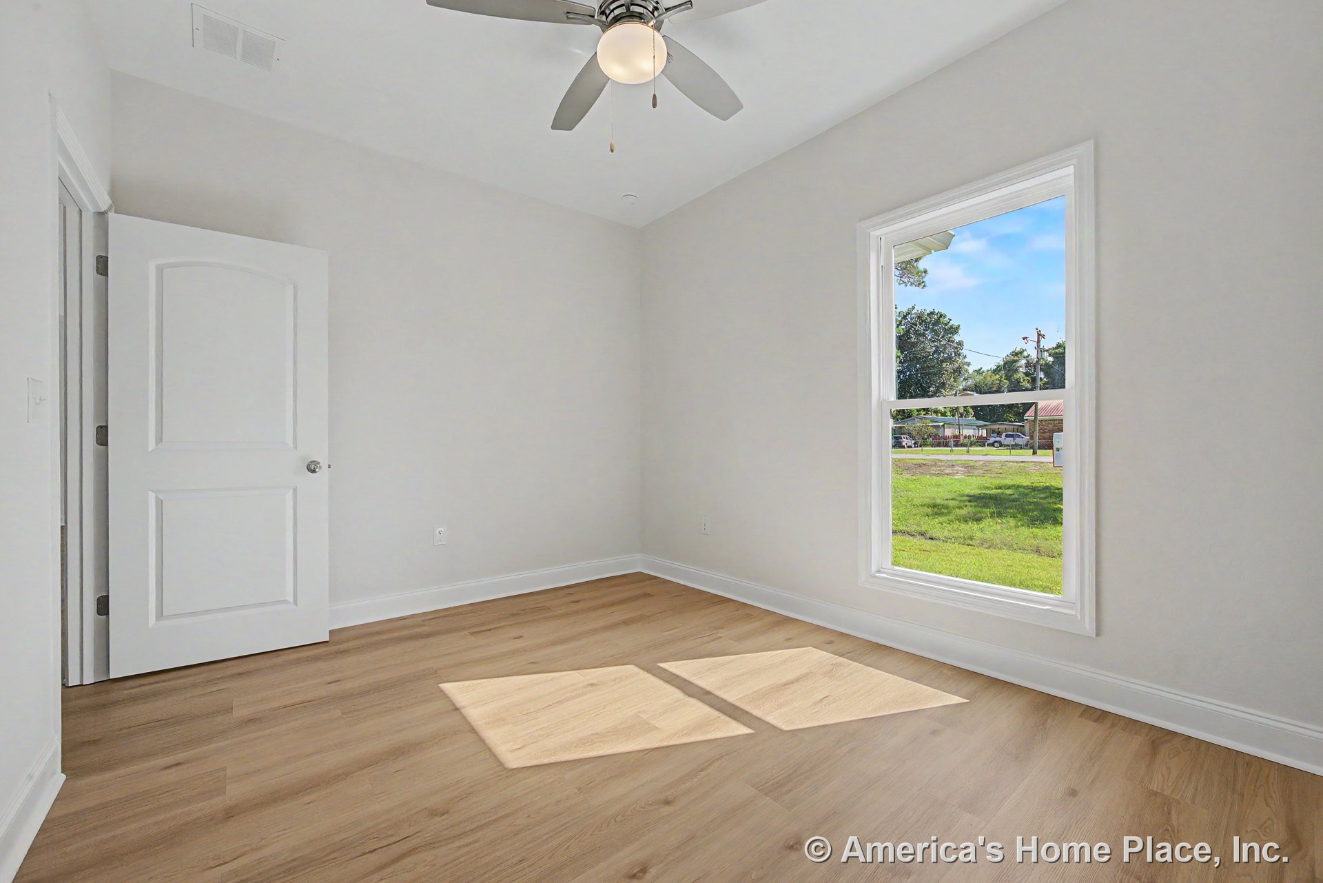 Light wood plank flooring, white trim and baseboards, large single window allowing natural light, ceiling fan with integrated light fixture, white paneled door, and neutral painted