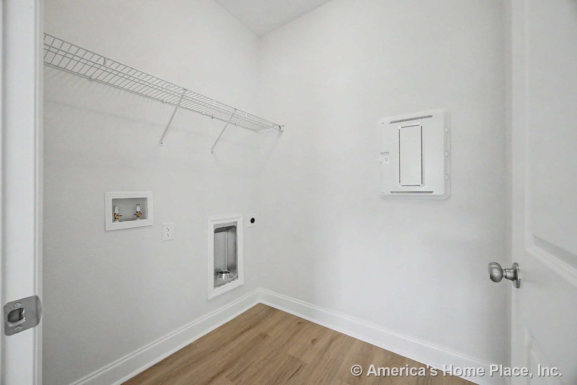 Laundry room with wall-mounted wire shelf above washer and dryer hookups, utility wall panel, white trim, light wood flooring, and white painted walls.