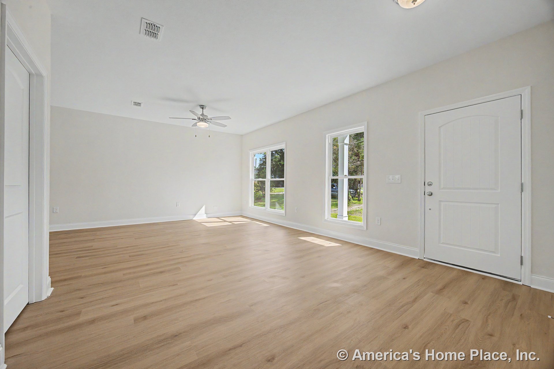 Light wood-look flooring, large double windows allowing natural light, white paneled front door, ceiling fan with built-in light, neutral wall color, and simple baseboard trim in a