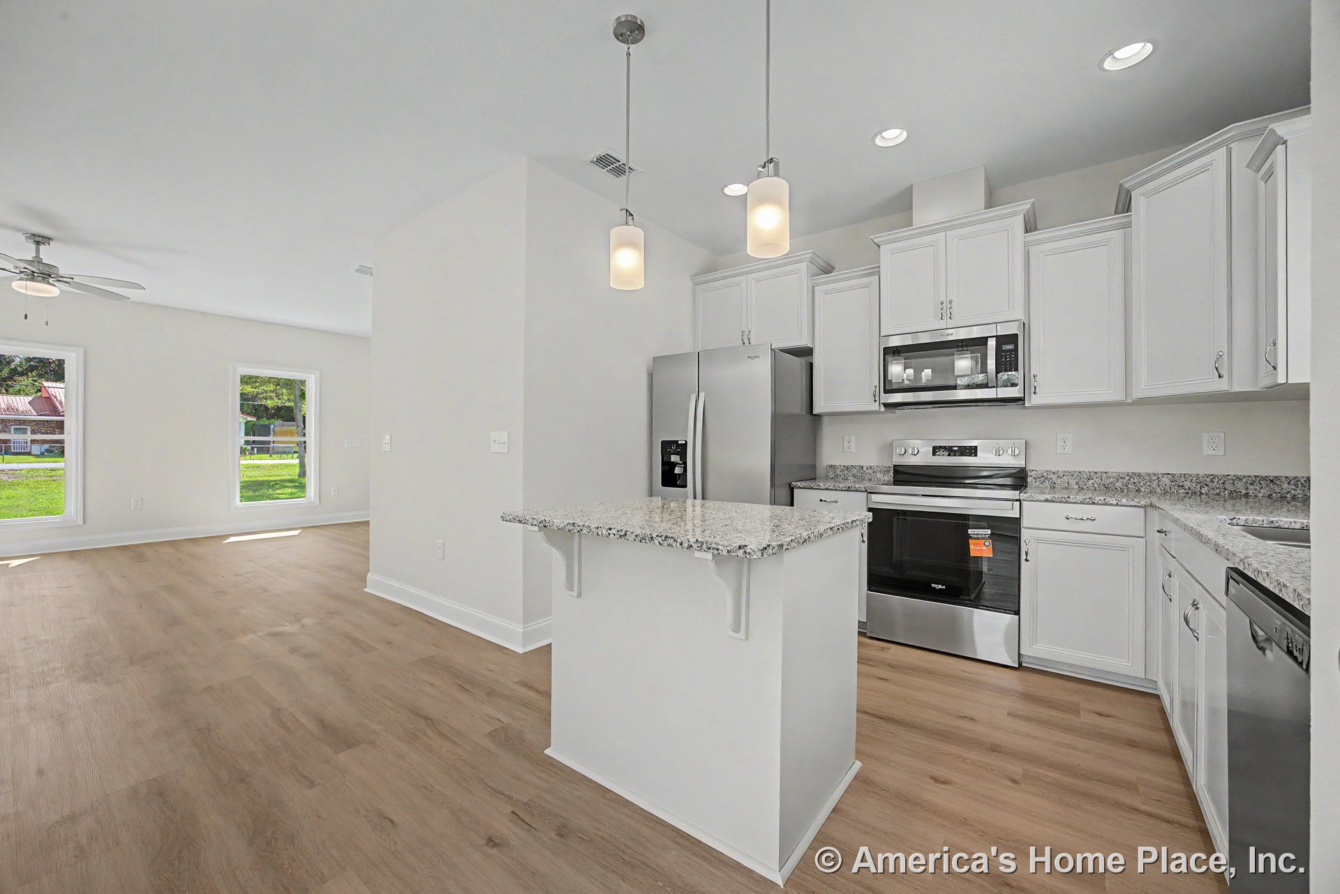 White kitchen cabinetry and granite island with stainless steel refrigerator, pendant lights, wood-look flooring, and open layout connecting to living area with large windows.