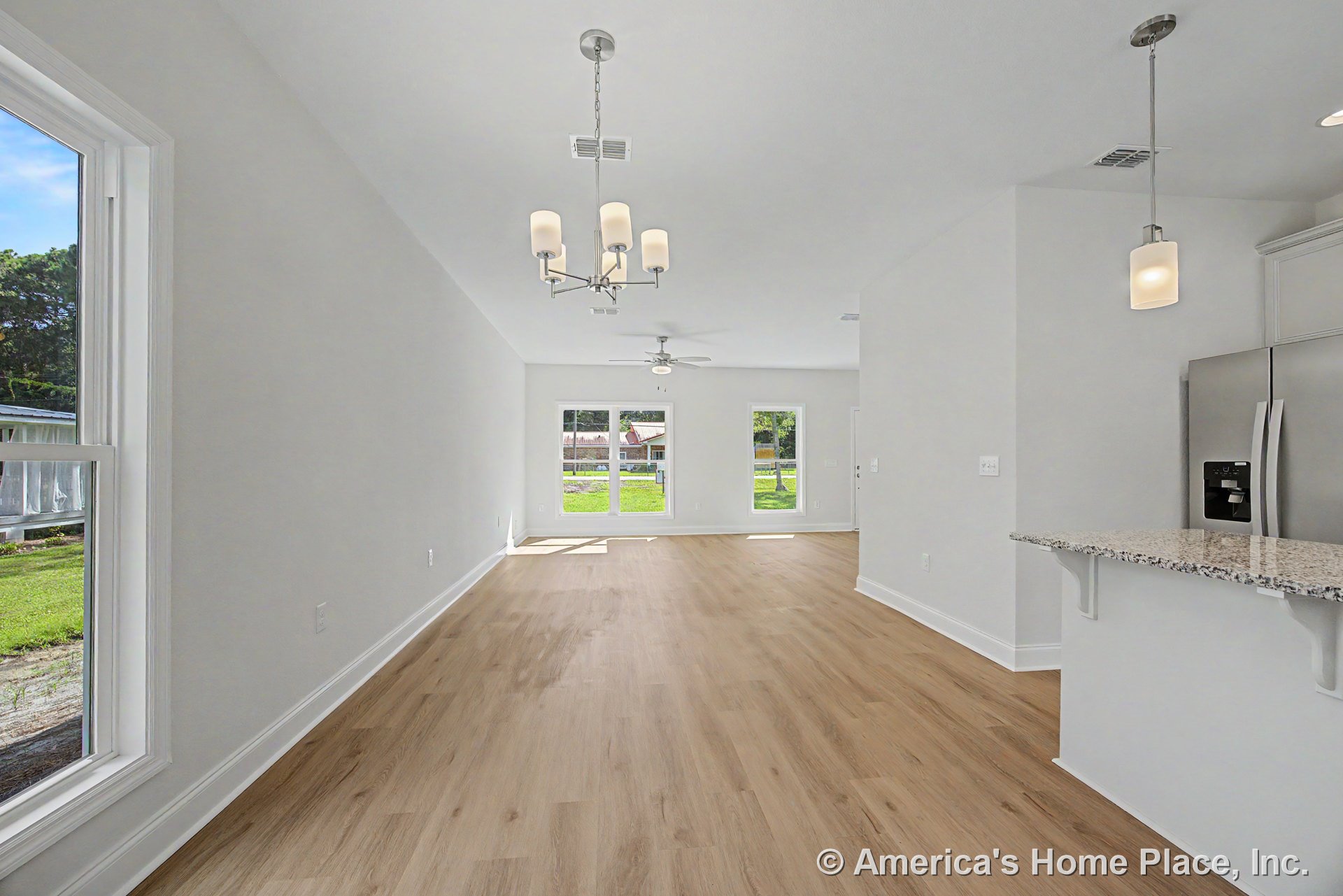 Open concept living and dining area with light wood plank flooring, large windows framed in white trim, granite kitchen peninsula, stainless steel refrigerator, modern pendant