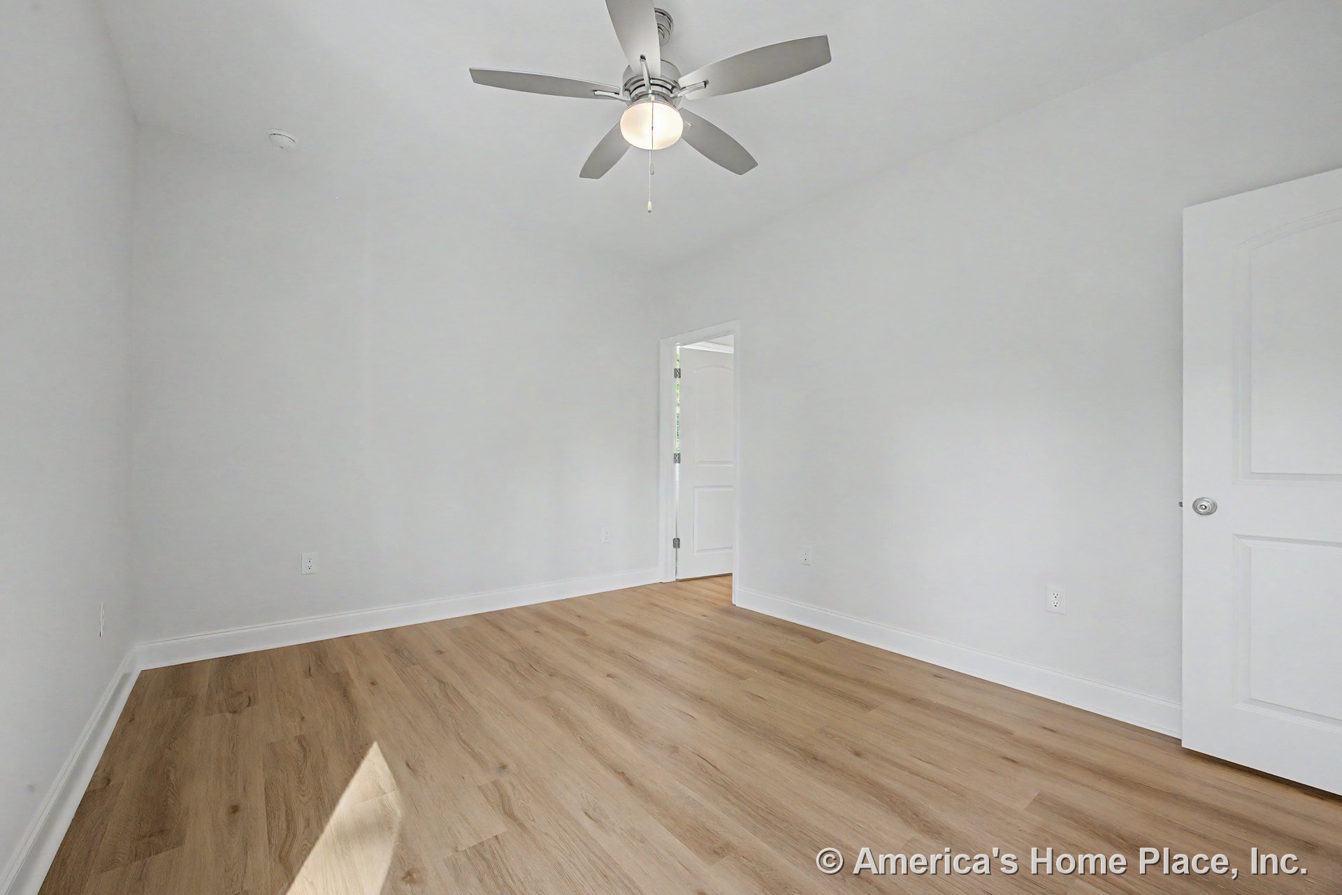 Light wood plank flooring, white baseboards, smooth white walls, white paneled door, ceiling fan with integrated light, single electrical outlet, minimalist modern finishes.