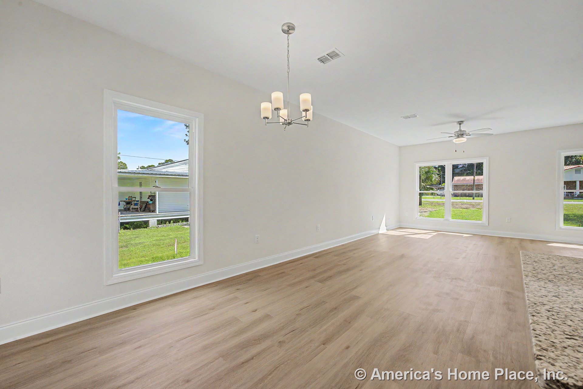 Open concept living and dining area with light wood plank flooring, large white-trimmed windows, granite kitchen countertop edge, modern chandelier, ceiling fan with integrated