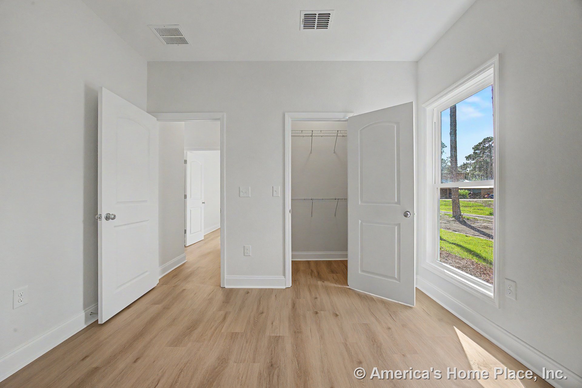 Bedroom with light wood plank flooring, large window with white trim, walk-in closet featuring wire shelving and white paneled doors, smooth white walls, ceiling vent, and