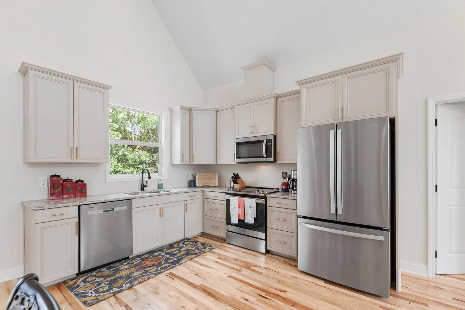 White kitchen with stainless steel refrigerator, microwave, and dishwasher, light cabinetry, stone countertops, oven with hanging towels, patterned rug on wood floor