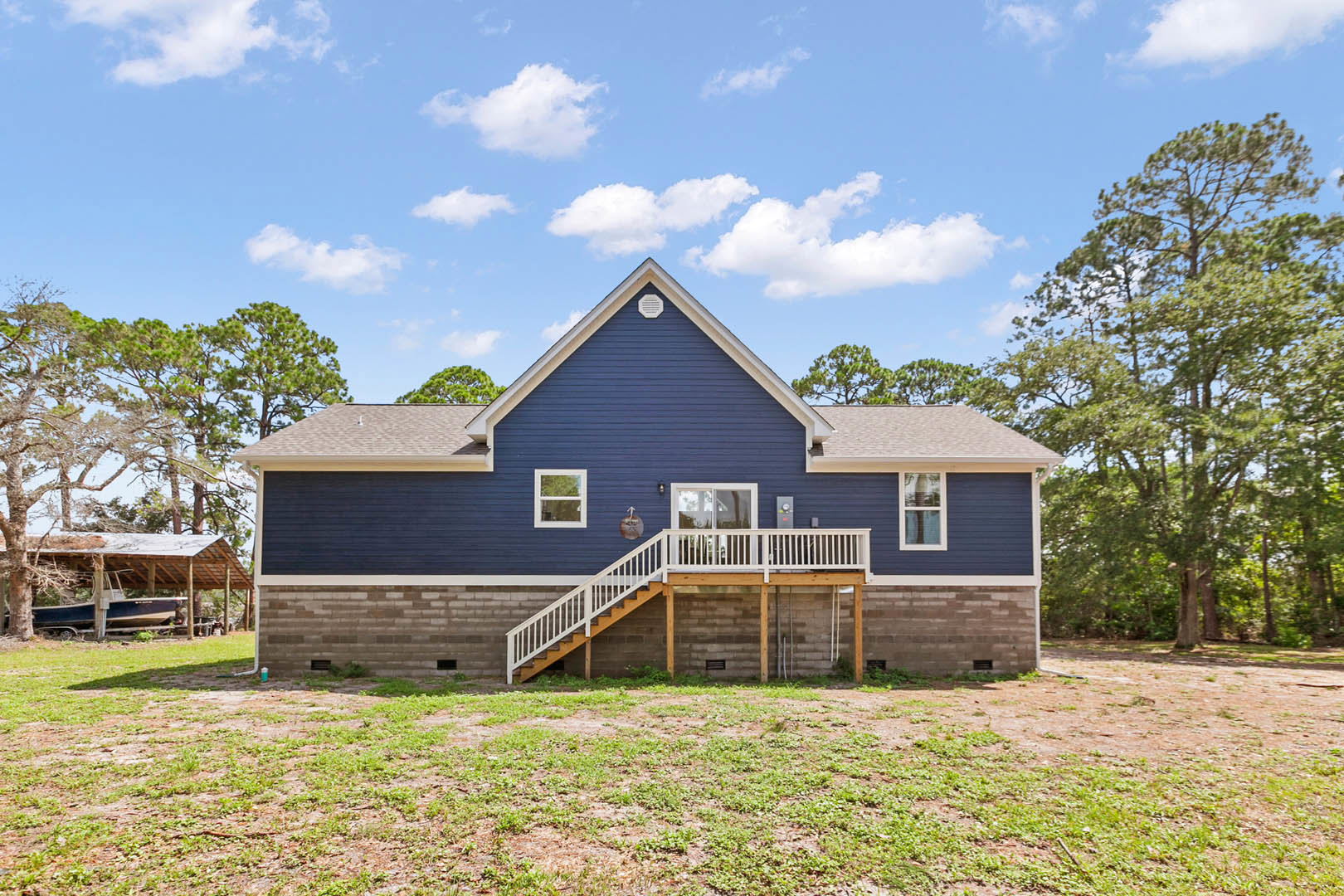 Two-story house with white framed windows, white porch railing, wooden deck and stairs, surrounded by green grass and mature trees under a partly cloudy sky