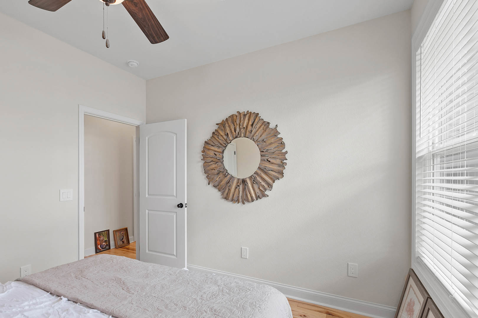 Circular wall mirror above white bed with matching bedspread, black door knob on white door, framed bird artwork, ceiling fan, window with white covering, plaster walls, small