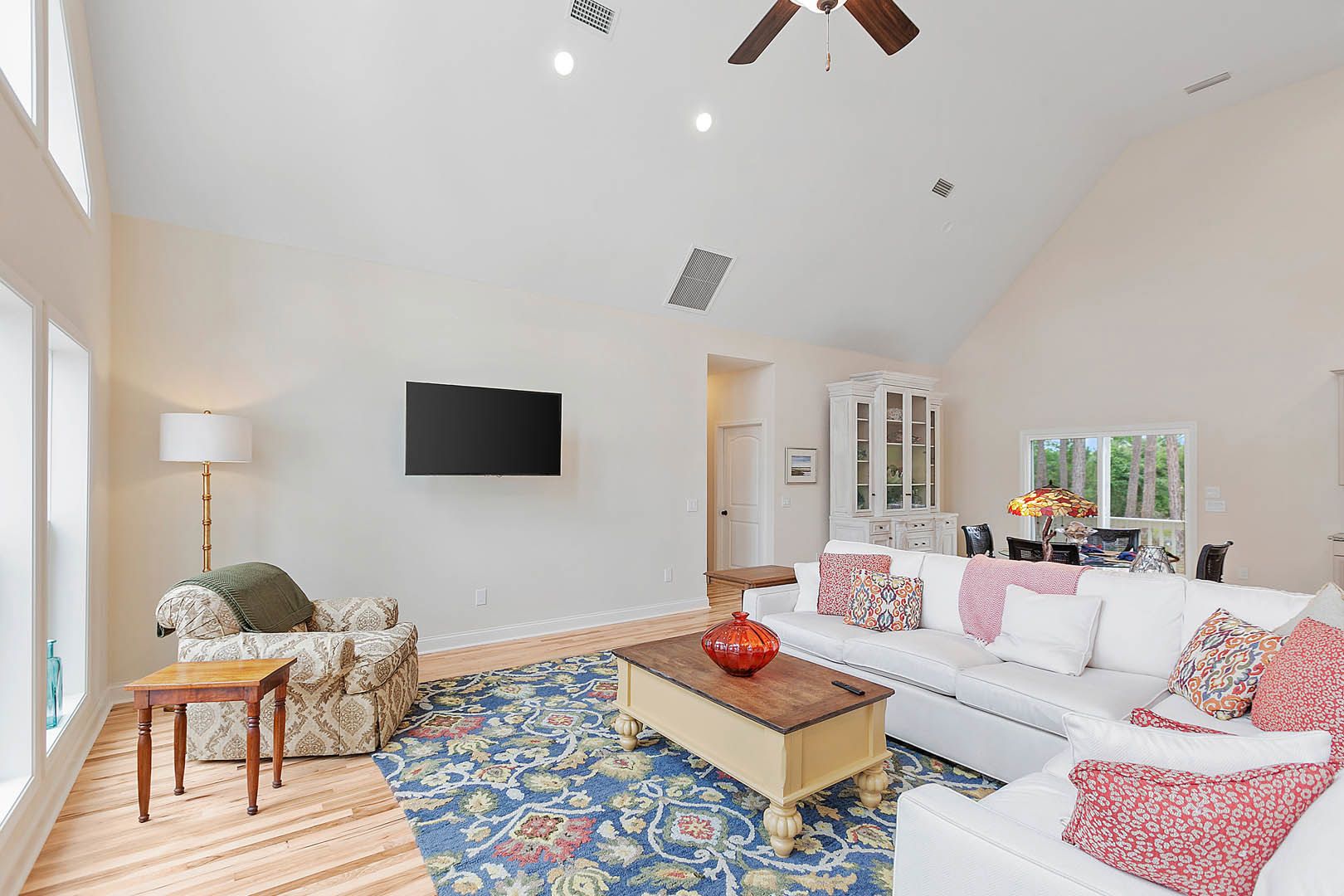 Spacious living room featuring a white couch with pillows, wooden table topped with a red vase, wall-mounted TV, and wood flooring