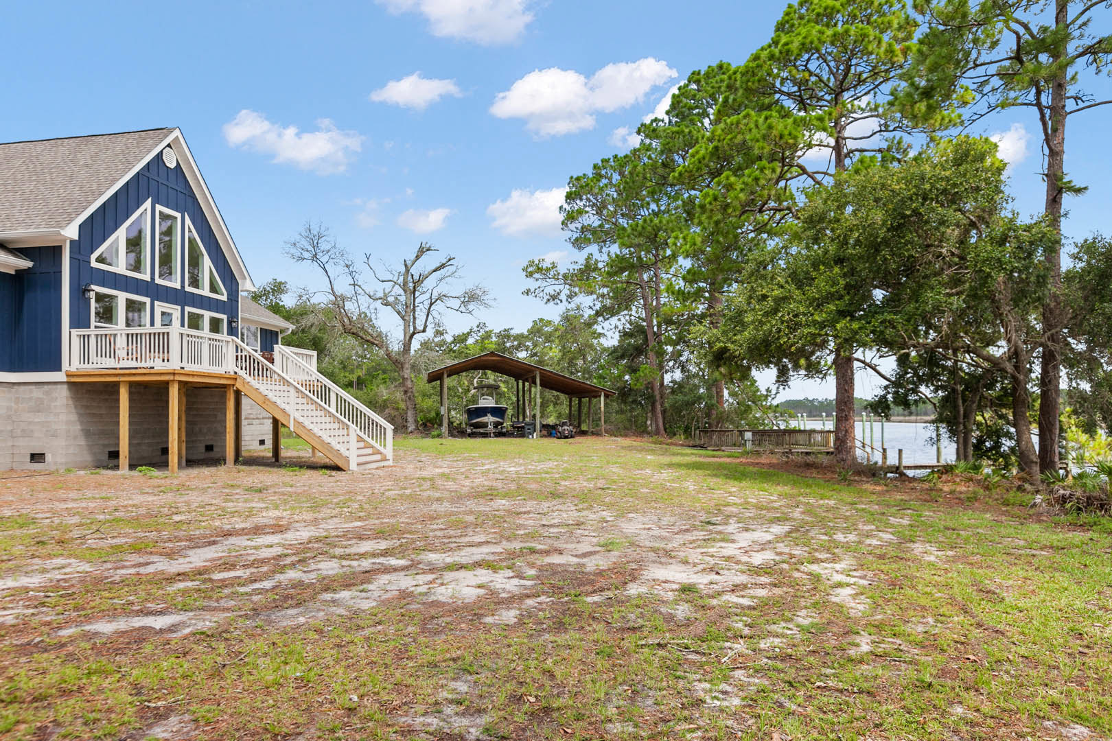 Two-story house with covered patio, white railing deck, stairs leading to grassy yard, surrounded by trees, overlooking lake with dock and boat