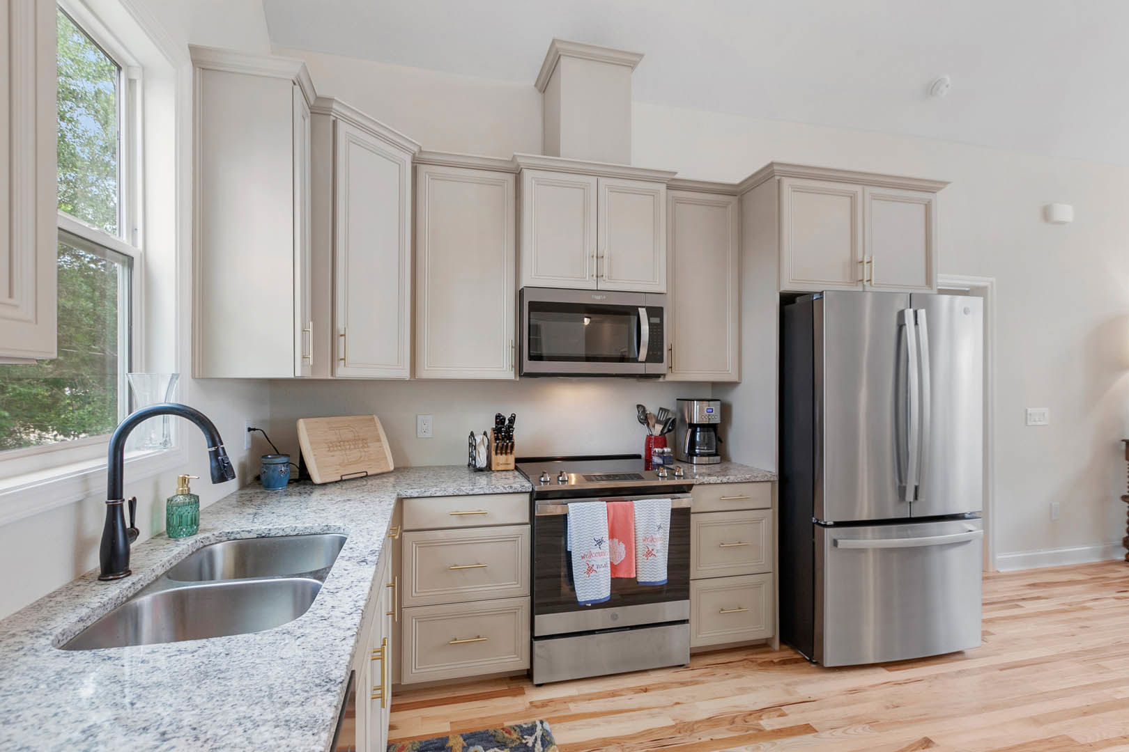 White kitchen cabinets, stainless steel refrigerator with handle, open microwave oven door, towels draped over oven, green bottle with gold top, wooden cutting board with carved