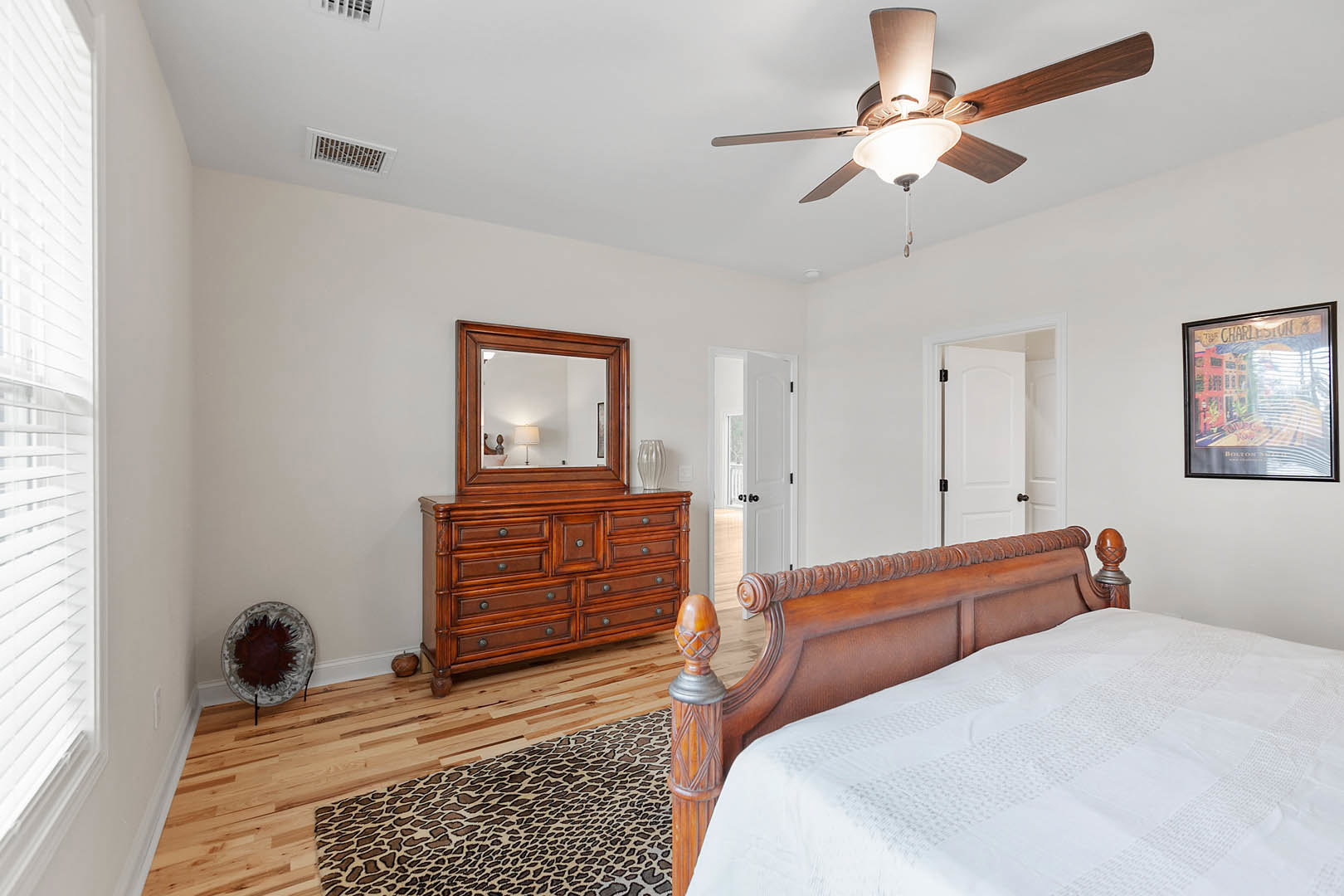 Bedroom with wood headboard bed, white bedding, dresser topped with mirror and decorative plate, framed city picture on wall, ceiling fan with light, window with close-up view