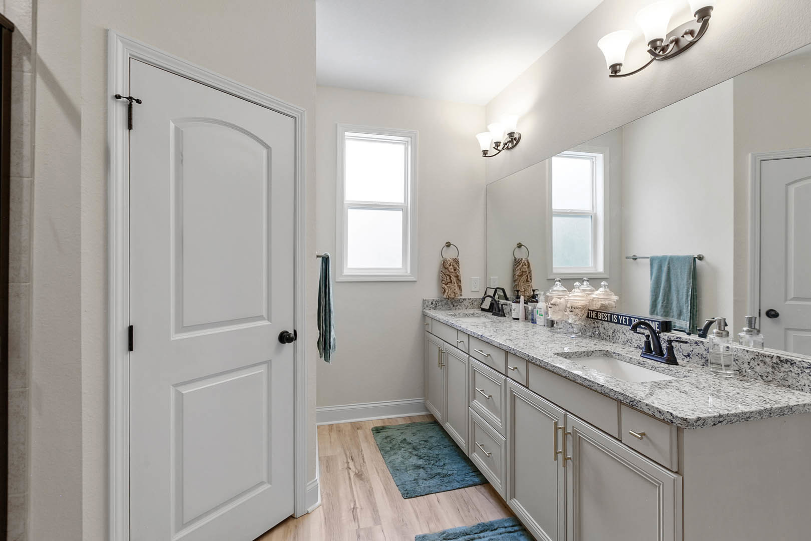 Bathroom with white shaker cabinets, blue area rug on tile floor, white door with black handles, towel hanging on metal ring, window with trim, modern light fixture above sink and