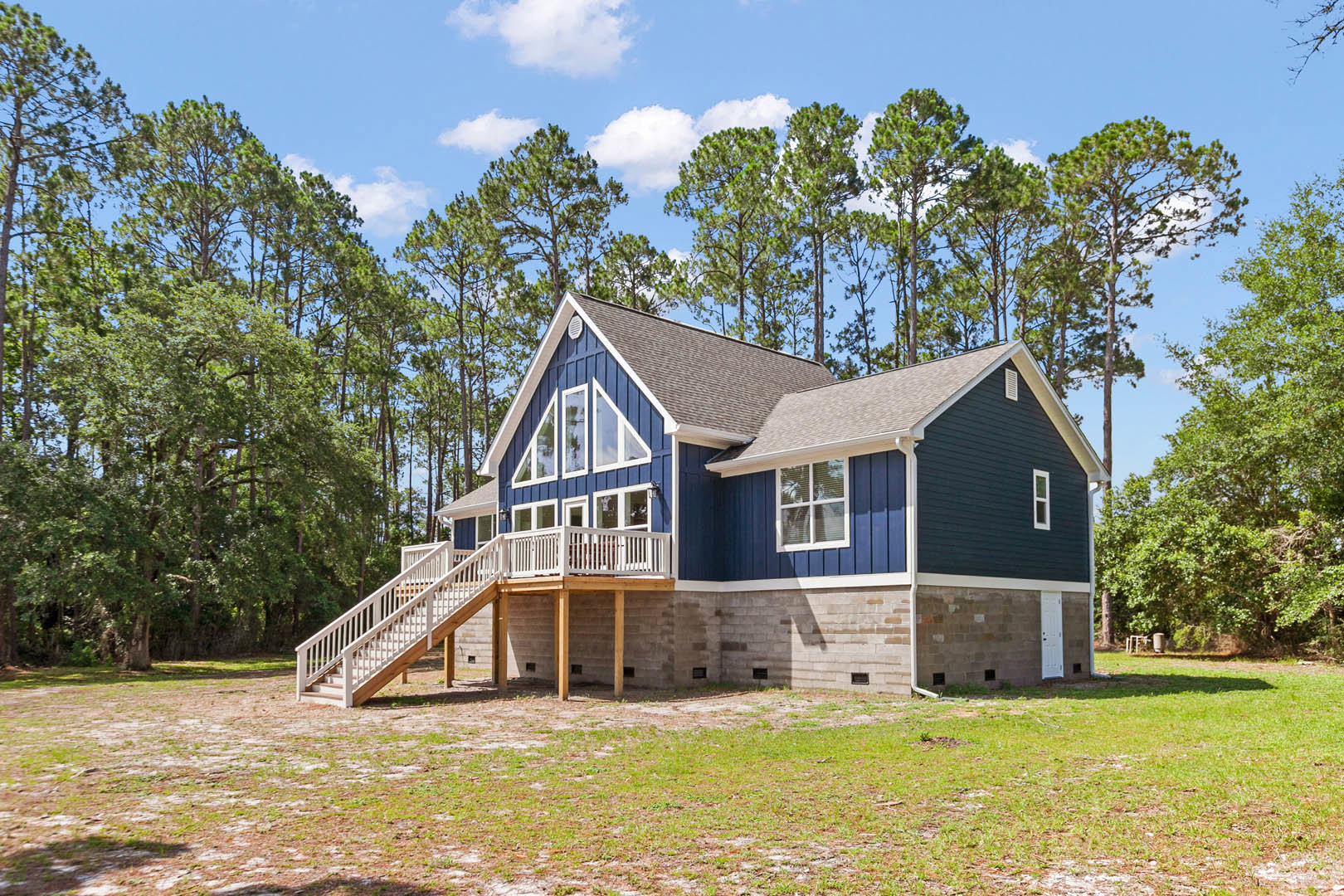 Blue house with wooden porch and stairs, white railing, window with closed blinds, grassy yard bordered by concrete wall, trees and clouds in background