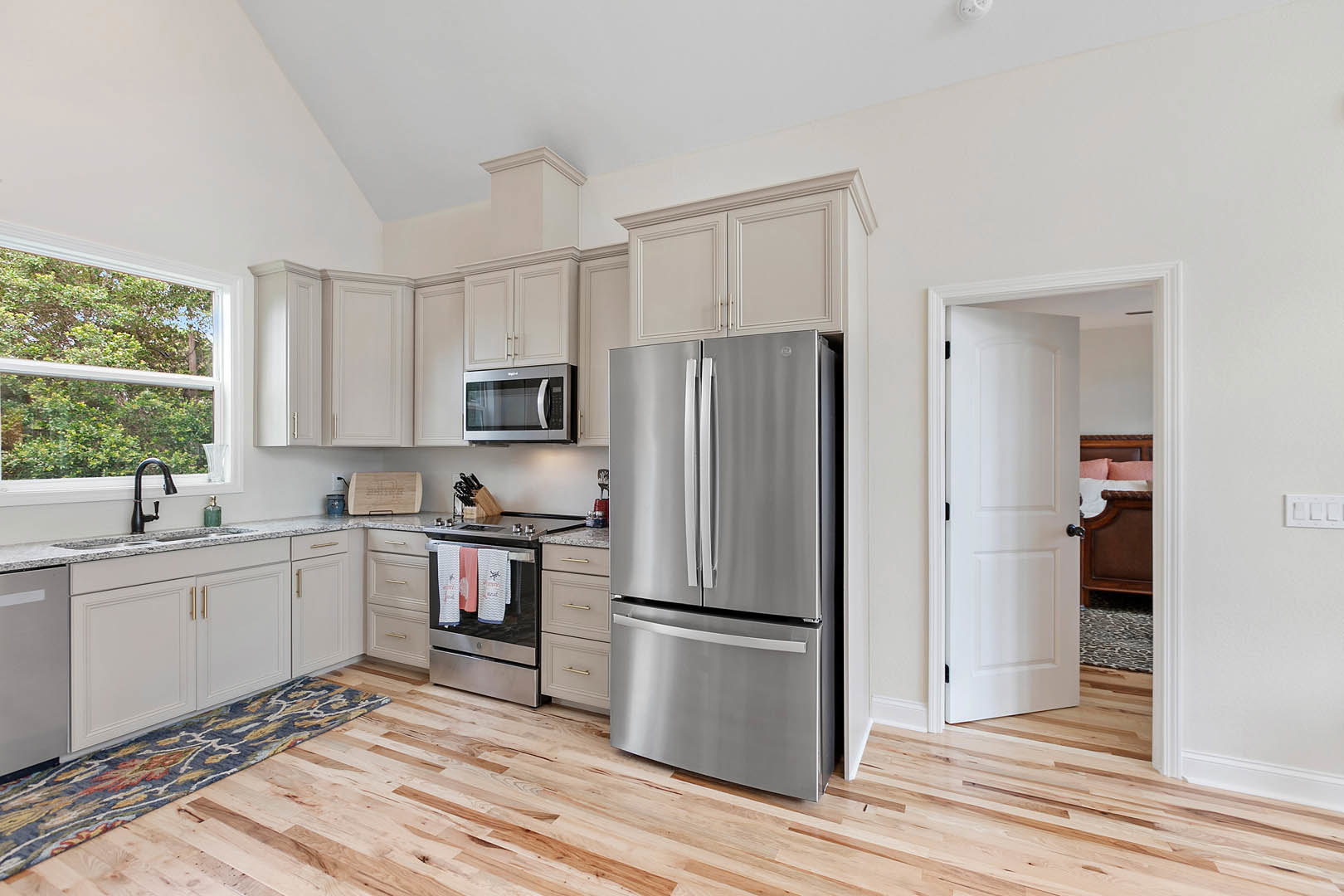 White kitchen cabinets, stainless refrigerator, open microwave, wood flooring, window above sink with a vase, oven with towels, white rectangular appliance with black border on