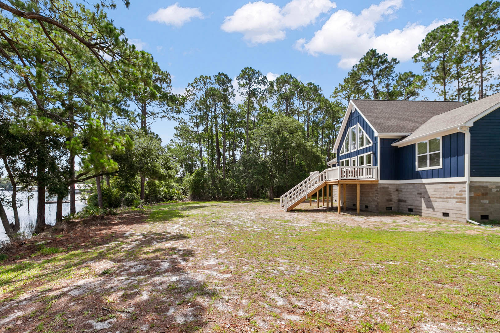 Blue siding house with white trim and railing, elevated deck overlooking grassy yard, staircase leading down, mature trees and blue sky in background