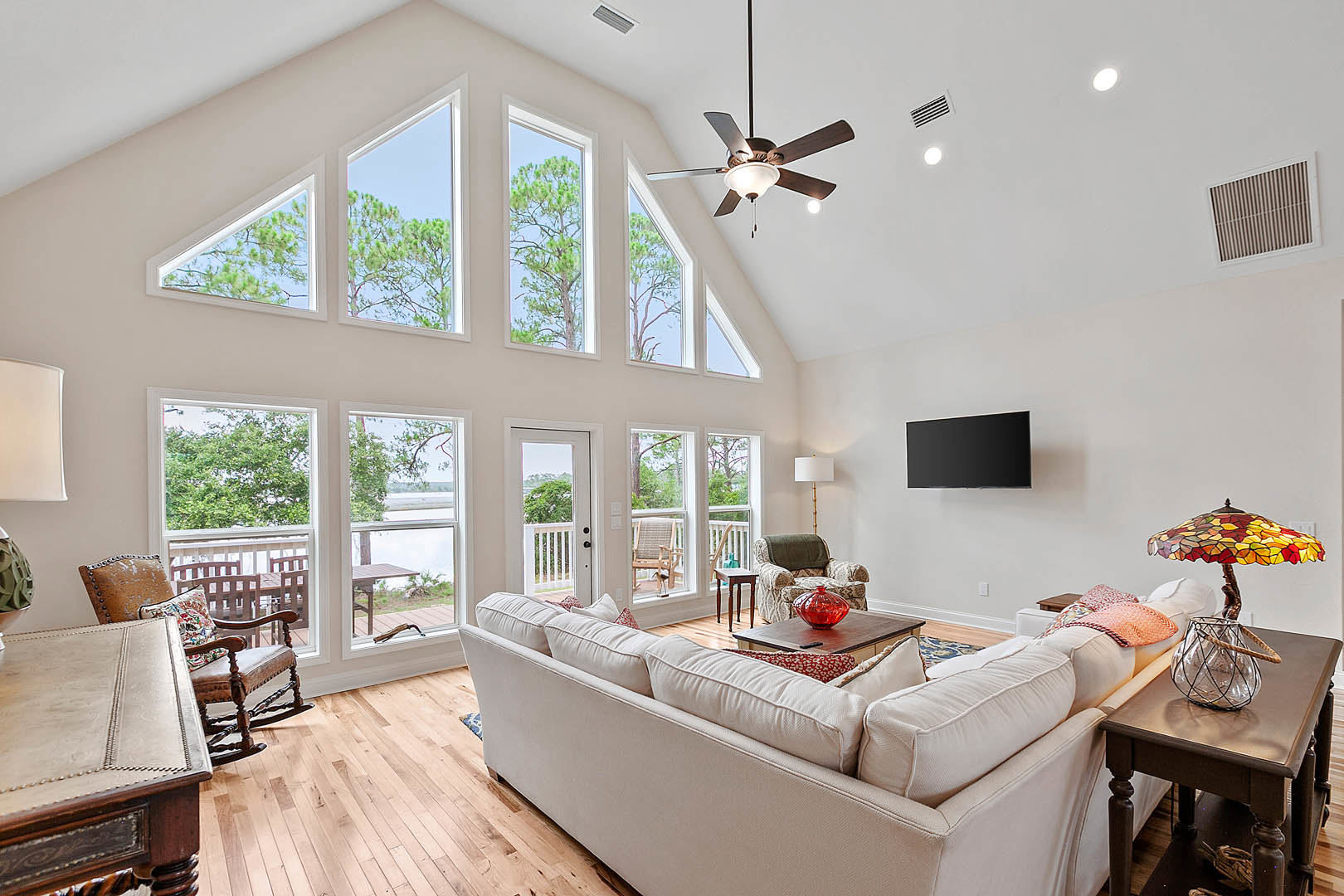 Living room with light wood flooring, gray sectional couch with pillows, glass coffee table holding a vase, chair draped with a blanket, large ceiling fan with integrated light