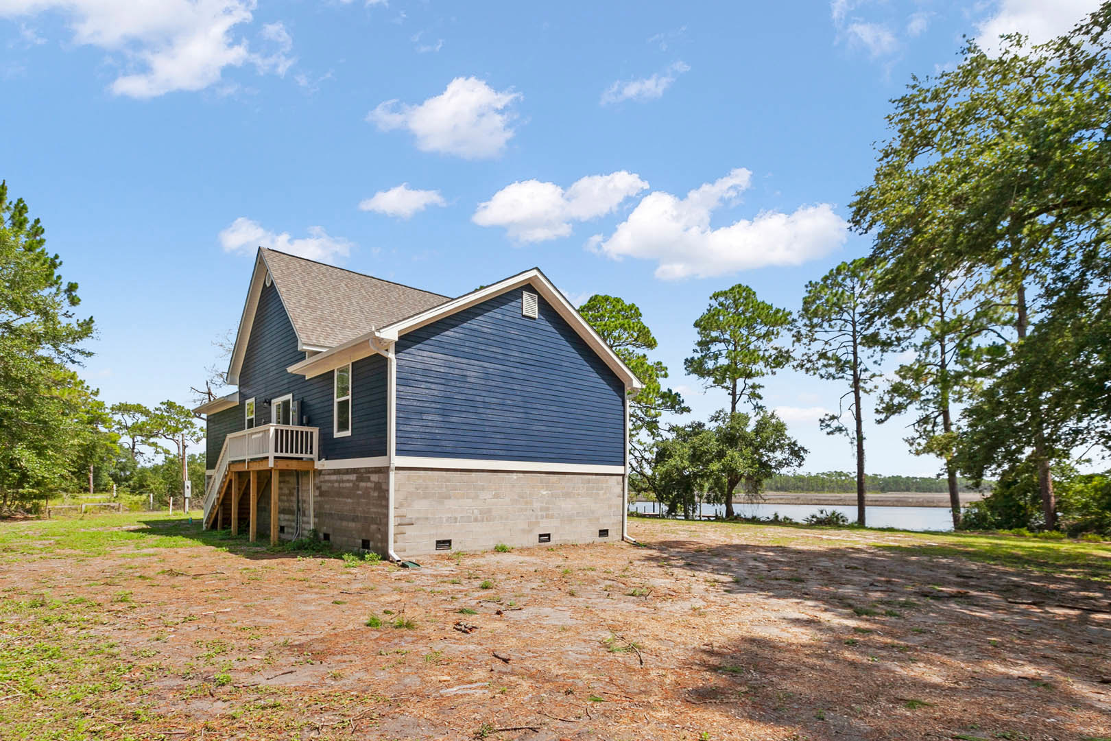 Blue-sided house with covered porch overlooking a lake, surrounded by trees and grass, brick retaining wall on dirt ground, gabled roof under clear blue sky