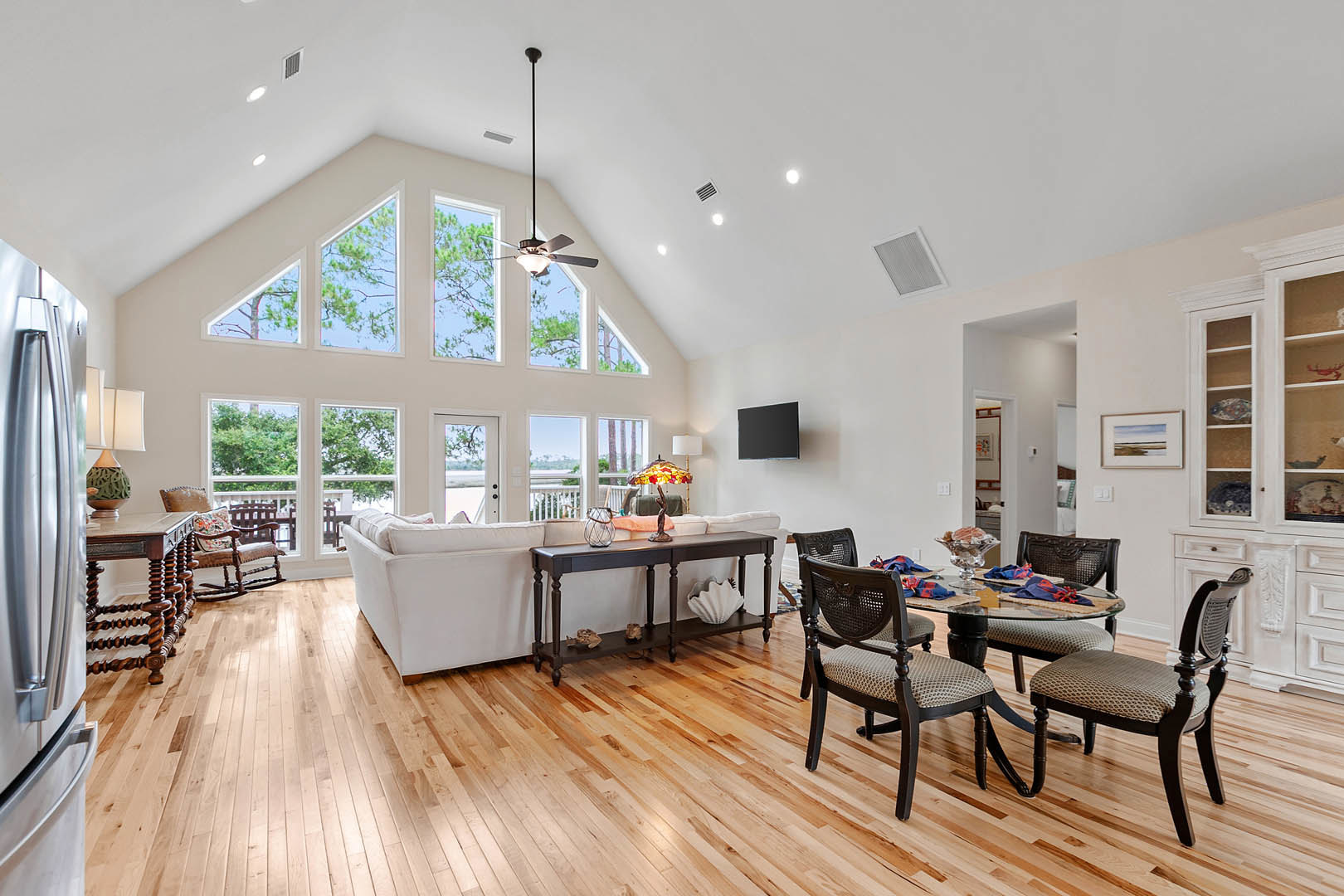 Living room with gray couch, upholstered chairs, wood coffee table, decorative vases on shelf, wall-mounted black TV, light wood flooring, white walls, and modern cabinetry