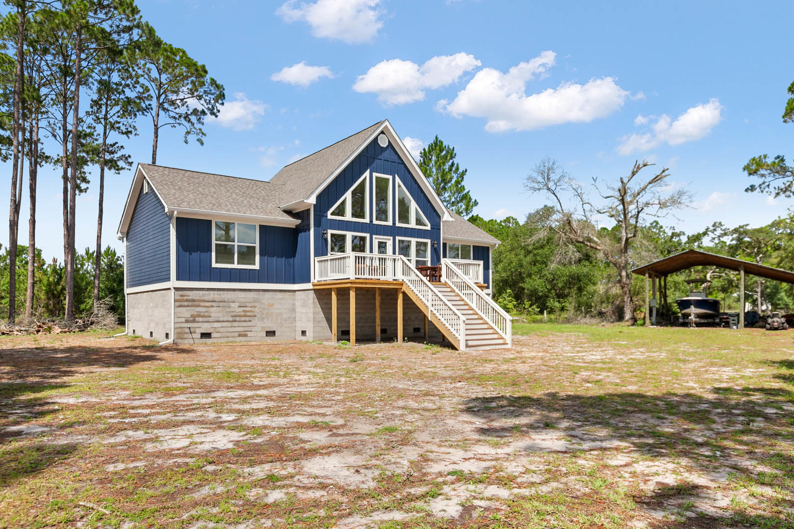 Blue cottage-style house with white trim, covered porch, wooden deck, fenced yard, leafy tree in background, patch of grass and dirt in foreground, partly cloudy sky overhead.