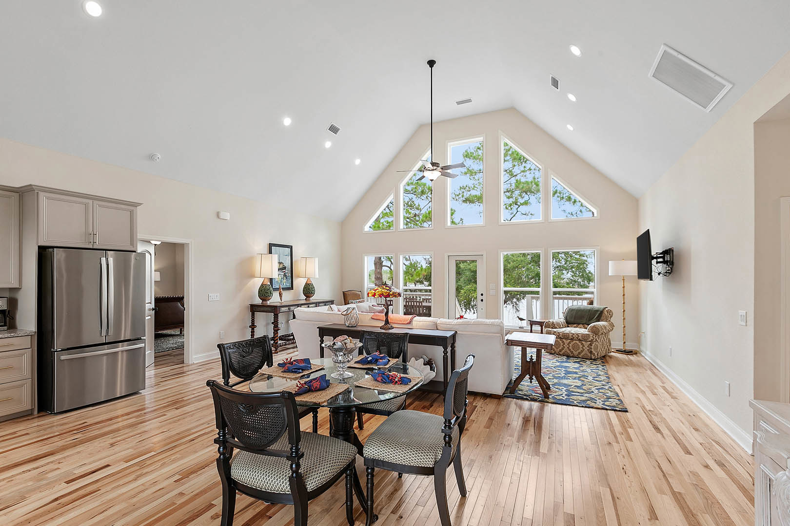 Open dining room with vaulted ceiling, glass-top table surrounded by wooden chairs, laminate wood flooring, and modern kitchen appliances including a stainless refrigerator.