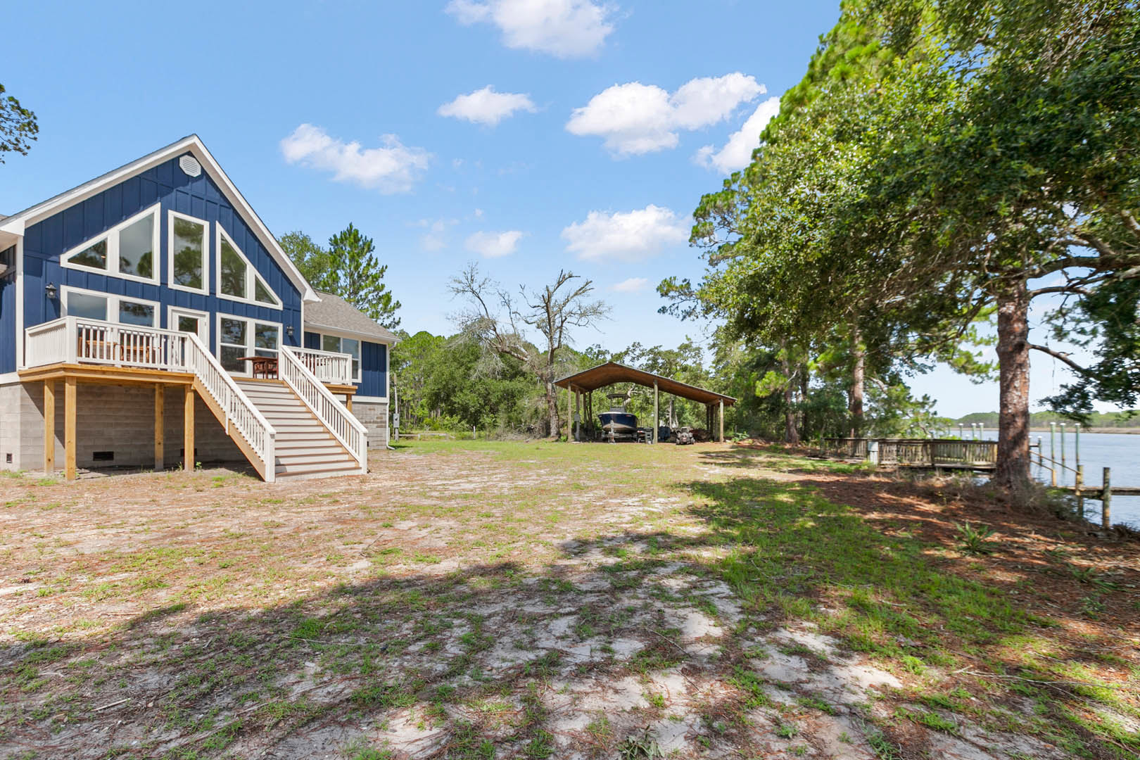 Covered porch with white railings, gray siding, and a car parked behind the house; mature trees and grass surround the property, with a wooden deck and outdoor seating visible.