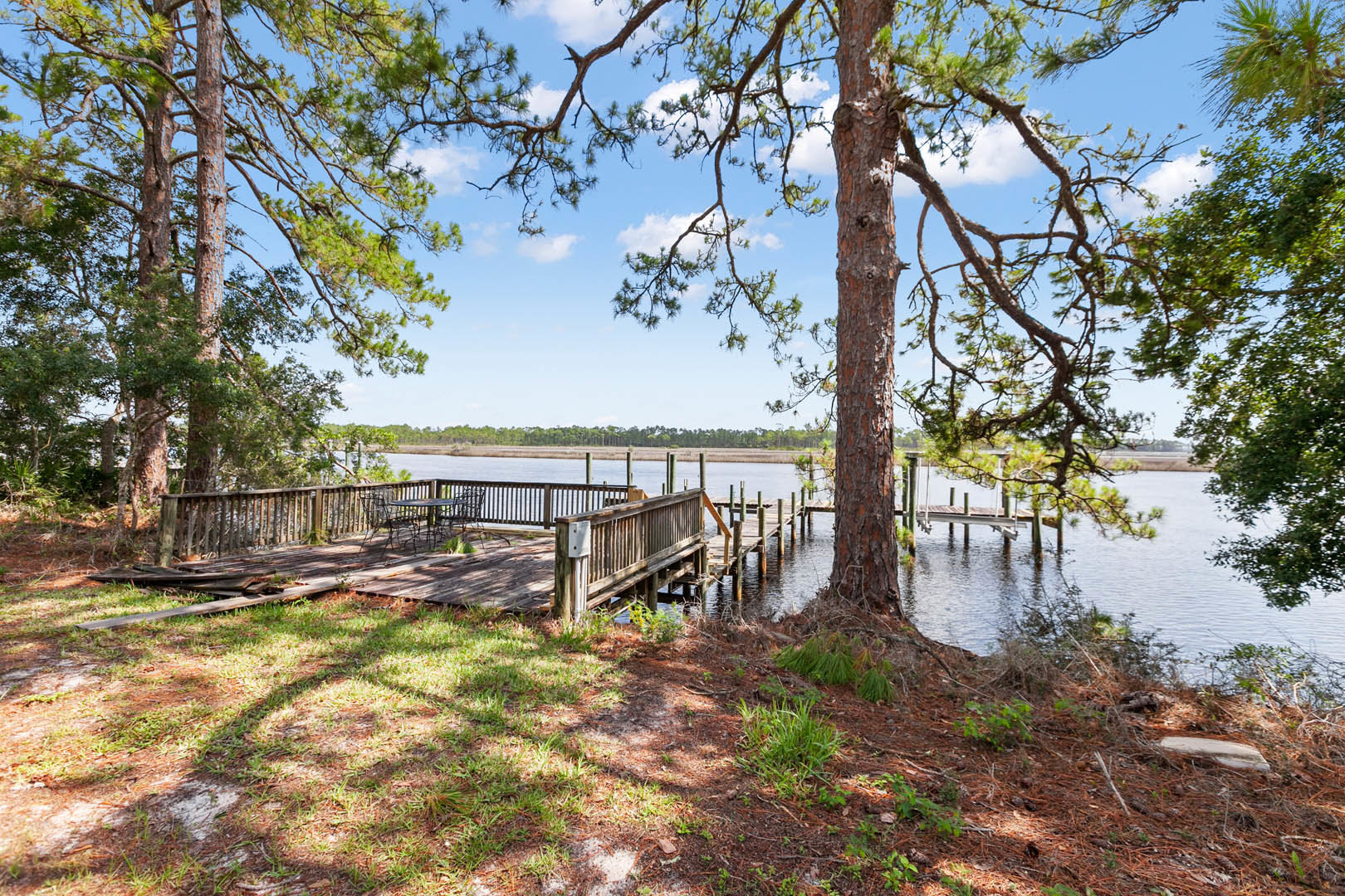 Wooden dock extending over calm lake, surrounded by lush grass and mature trees, outdoor deck with table and chairs visible, partly cloudy sky reflected in water.