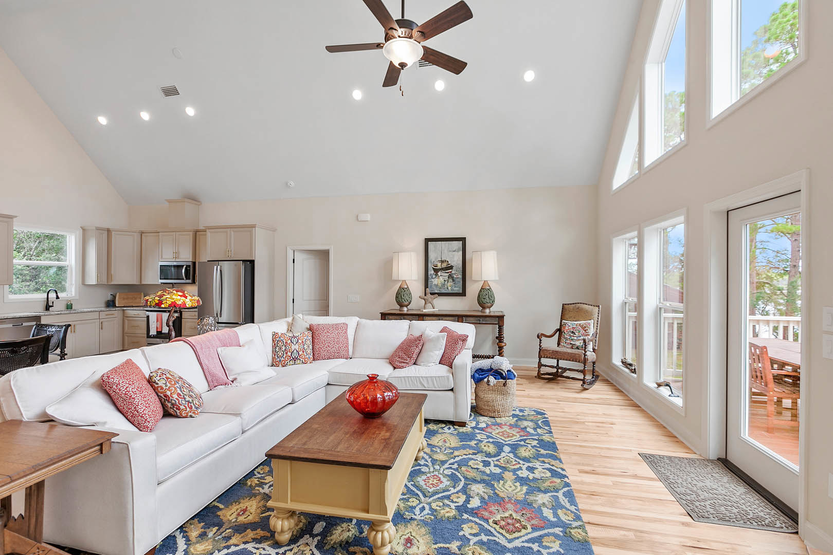 Spacious living room featuring a white couch with pillows, a wooden table topped with a red glass vase, a chair with a pillow, neutral walls, and a ceiling fan with light fixture.