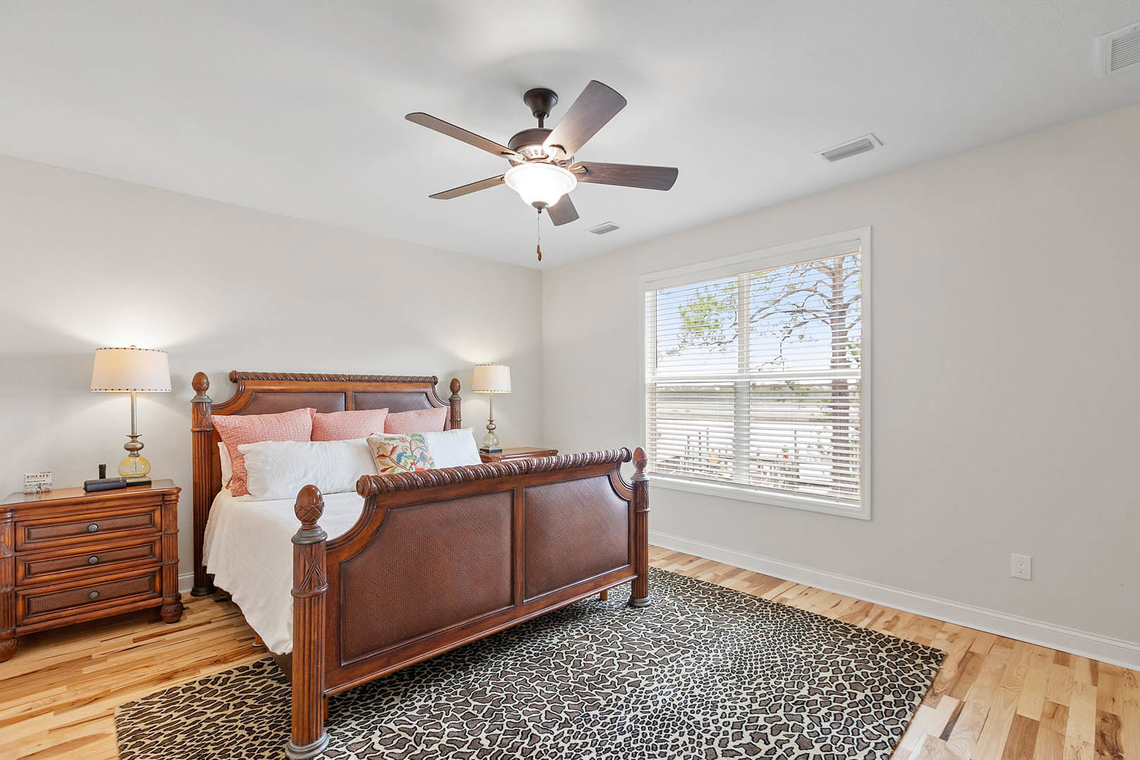 Bedroom with a wooden dresser, bed with white linens, bedside lamp with white shade, ceiling fan with light, window with white blinds, neutral walls and flooring