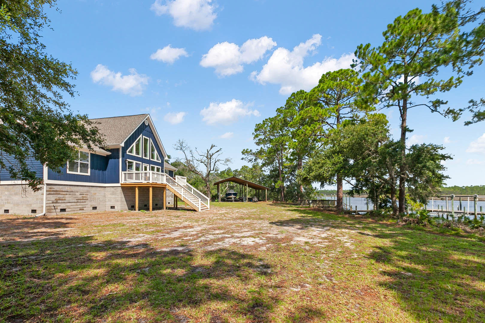 Blue cottage-style house with white railing, surrounded by mature trees and expansive grassy yard, staircase leading toward a boat under a canopy near a lake.