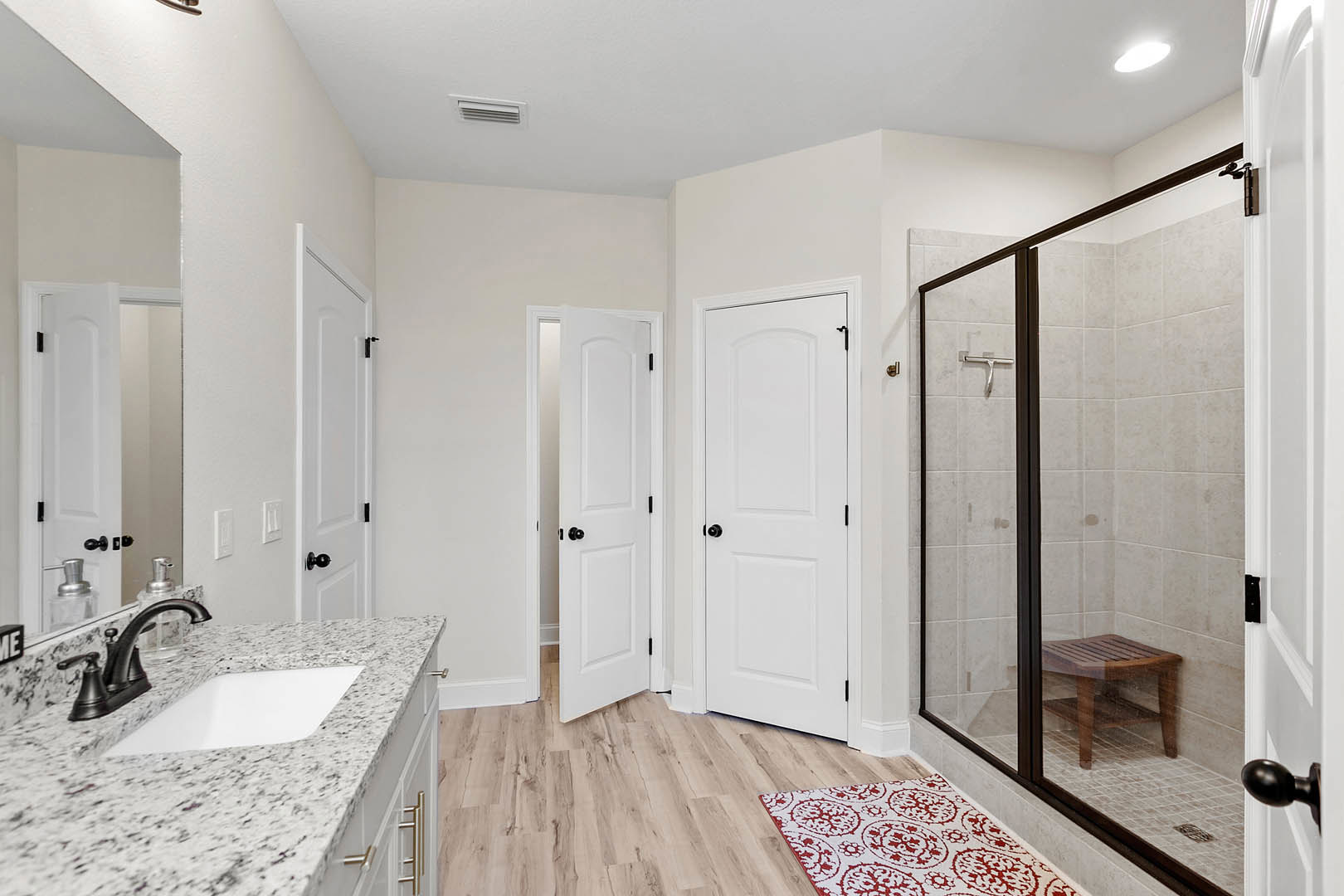 Bathroom with glass shower enclosure, white door with black knobs, red and white rug on wood floor, countertop sink with chrome faucet, wooden stool in corner, tile walls and